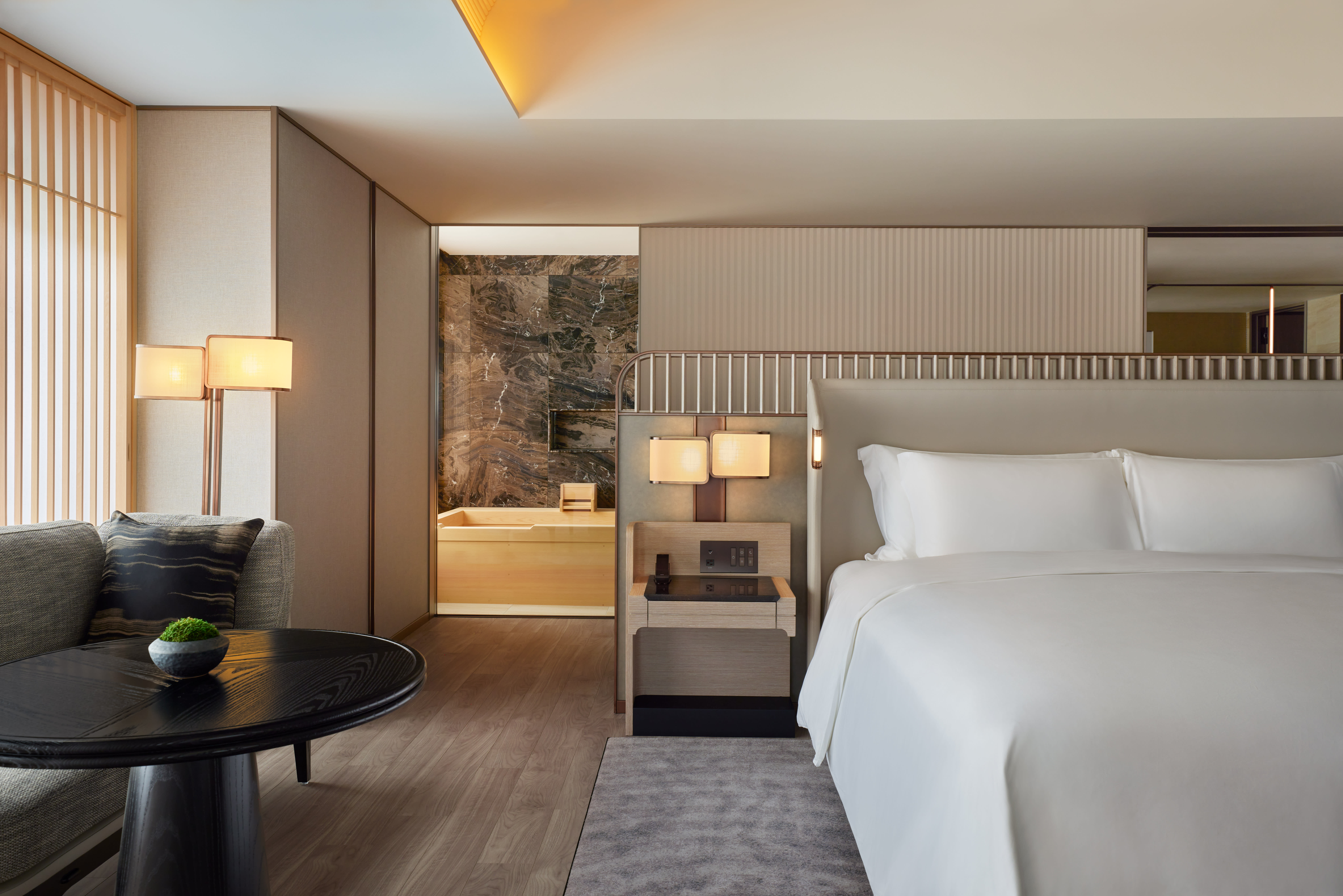 Modern hotel suite with a large white bed, soft neutral tones, and warm lighting. A cozy seating area sits to the left, while an open doorway reveals a bathroom with dark marble walls and a light wood soaking tub.
