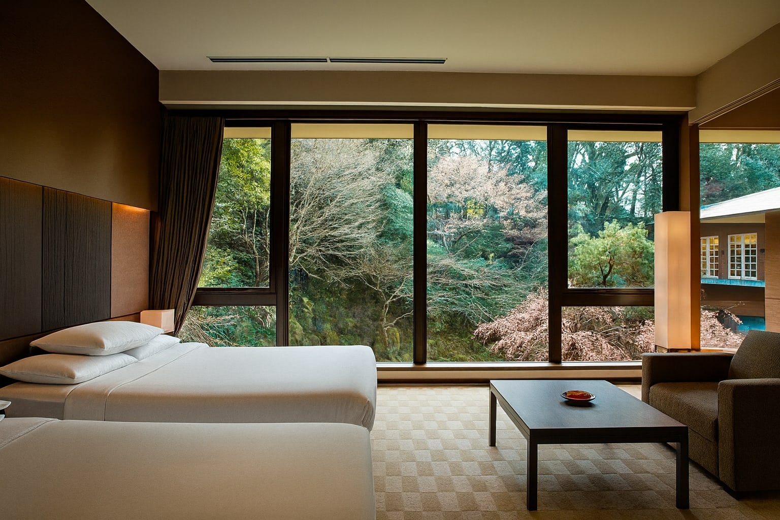 Spacious hotel room with two white beds facing a floor-to-ceiling window that overlooks a lush garden with trees and seasonal foliage.