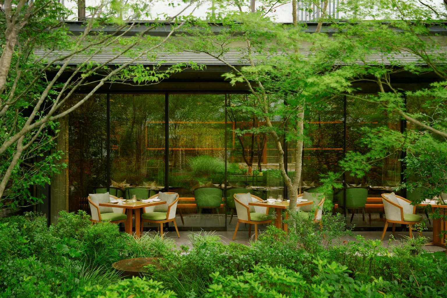 Outdoor dining tables and chairs sit beside floor-to-ceiling windows, surrounded by lush green foliage and leafy trees in a tranquil garden setting
