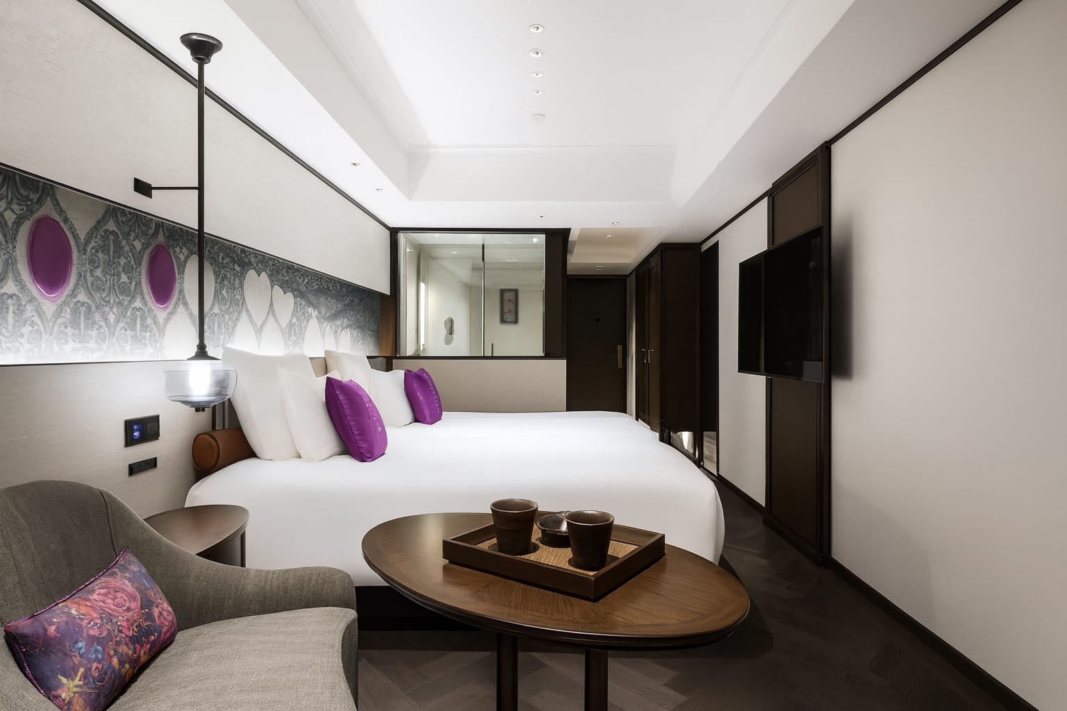 Contemporary hotel room with two white beds accented by purple pillows and a patterned headboard. A cozy seating area with a grey armchair and small wooden table sits in the foreground, while a large TV is mounted on the opposite wall.
