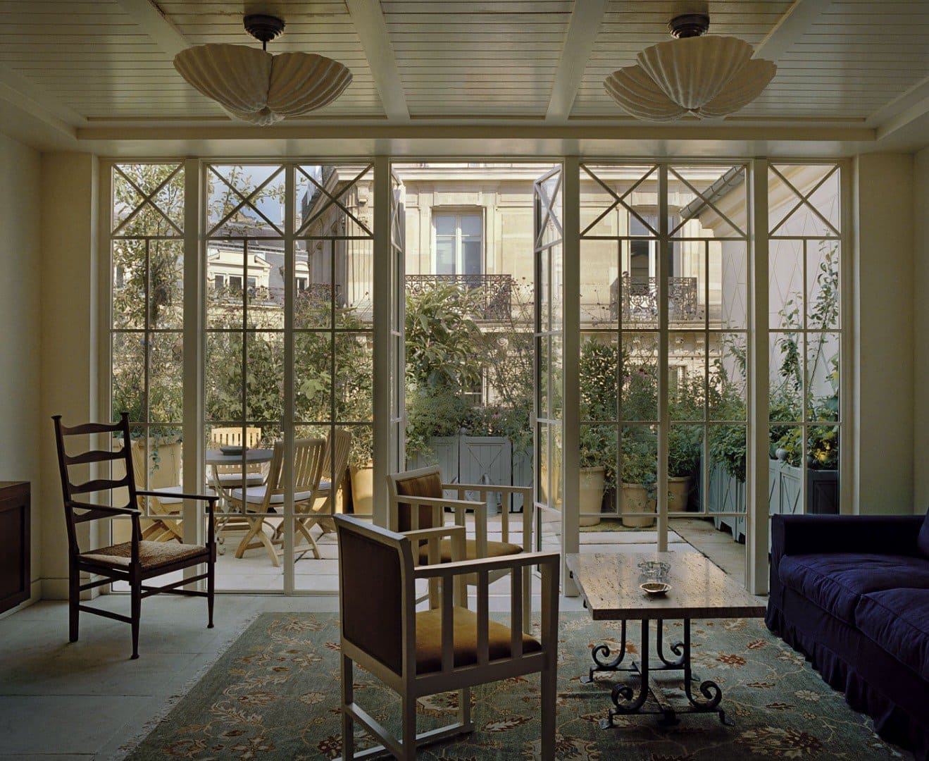 A sunlit sitting room with vintage-style armchairs, a sofa, and a wooden coffee table opens onto a terrace filled with potted greenery, framed by large grid-pane windows overlooking elegant Parisian architecture.