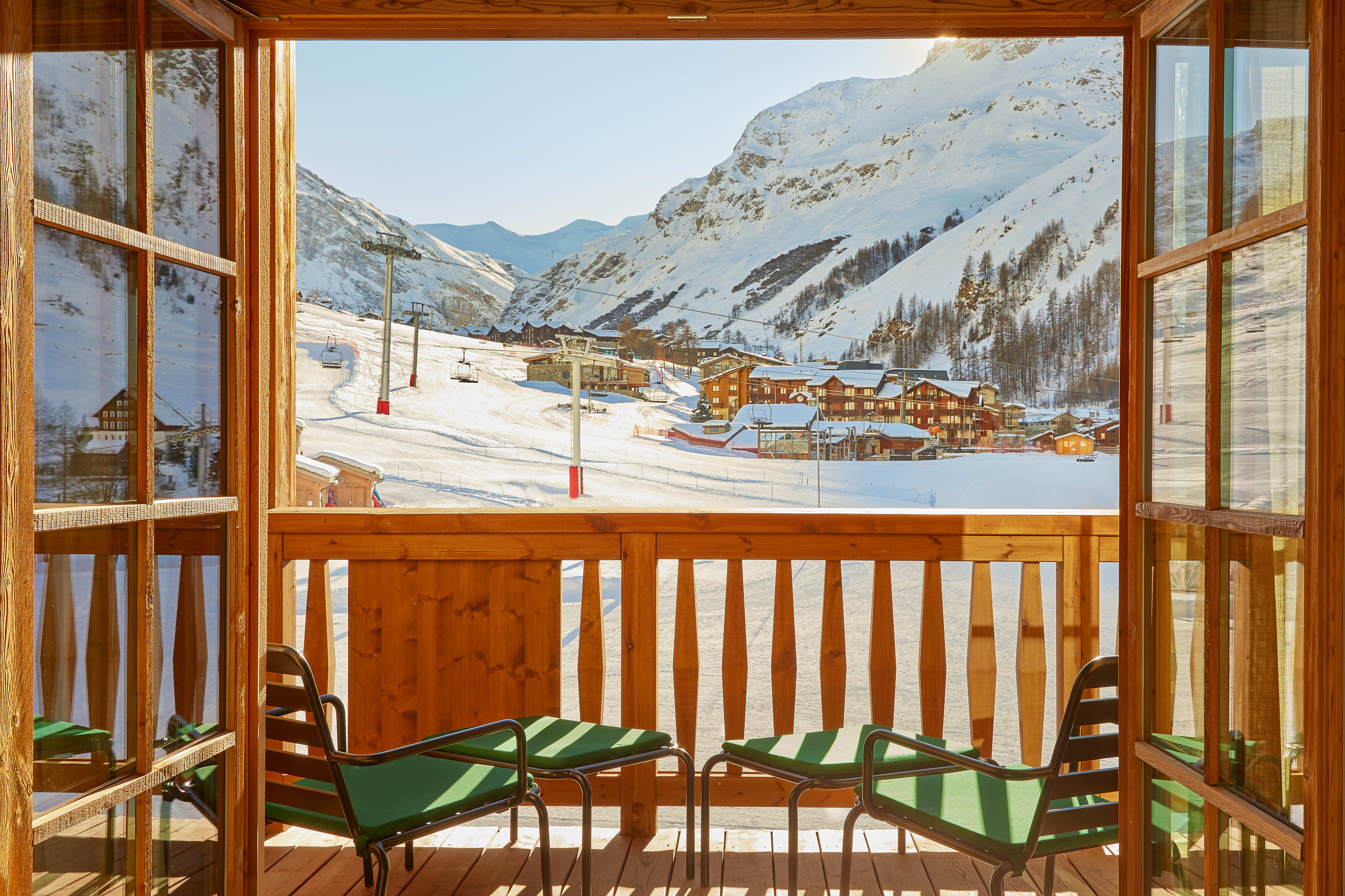 A wooden balcony with green chairs overlooking a snowy mountain ski resort, with ski lifts, chalets, and sunlit slopes framed by open wooden doors.