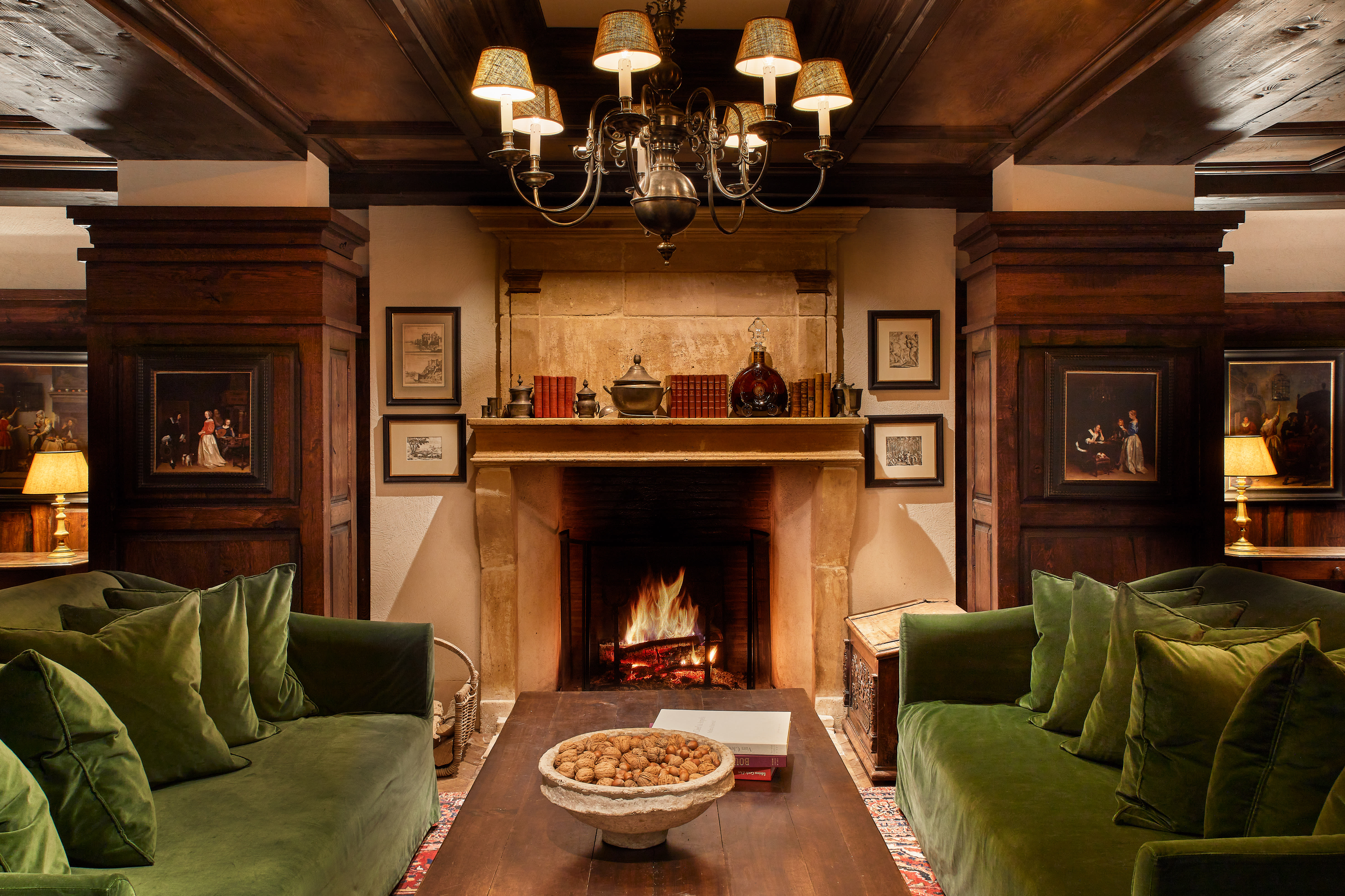 A cosy wood-panelled lounge with two green velvet sofas facing a stone fireplace, lit with a burning fire, framed artwork on the walls, a chandelier overhead, and a wooden coffee table topped with a bowl of nuts and books.