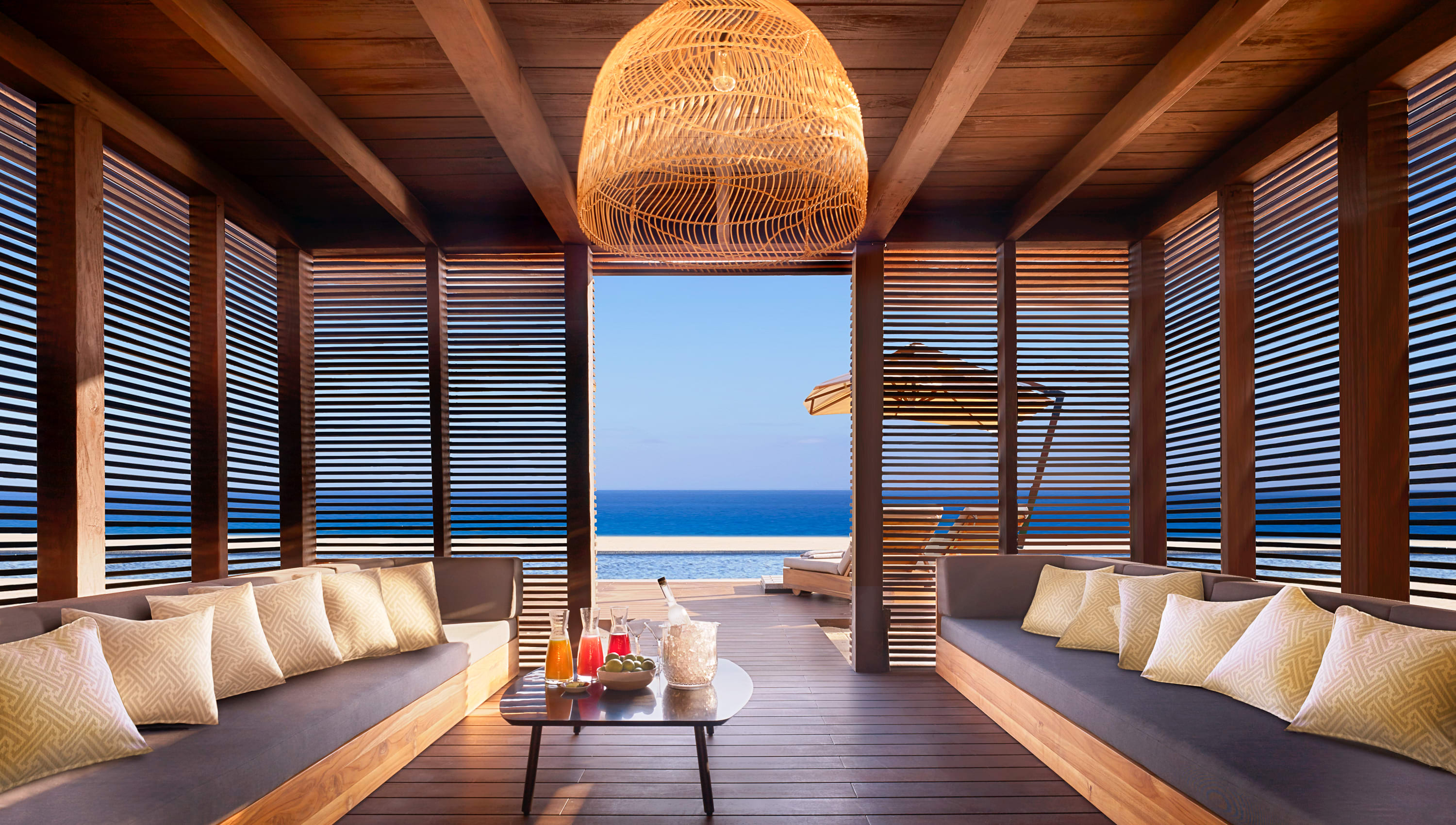 pen-air beachfront cabana with warm wooden beams, woven pendant light, and cushioned lounge seating, opening directly onto a sunlit deck with ocean views and the deep blue sea beyond.