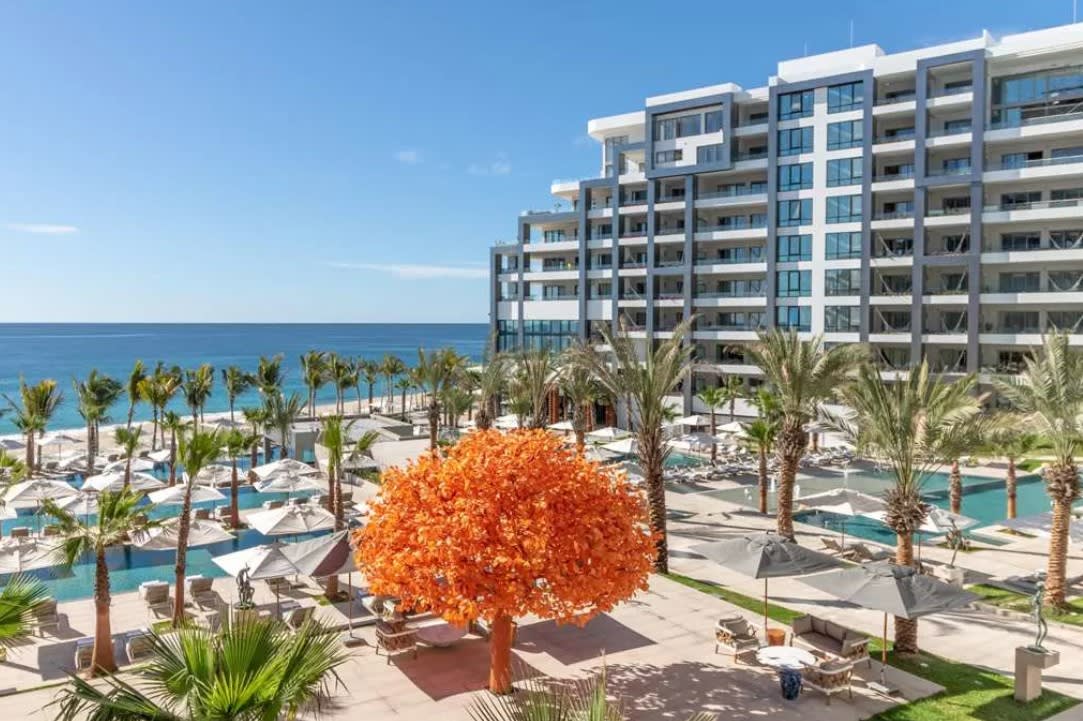 Modern beachfront resort with tiered white-and-glass buildings overlooking palm-lined pools and sun loungers, set directly on a sandy beach with calm blue ocean views stretching to the horizon.