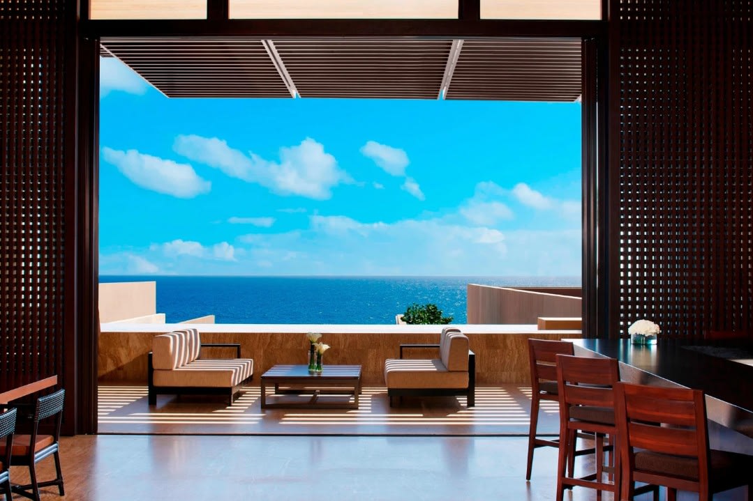 Open-air lounge with modern seating framed by wooden screens, looking out over a bright blue ocean and sky, with shaded sofas and a low table positioned to capture uninterrupted sea views.