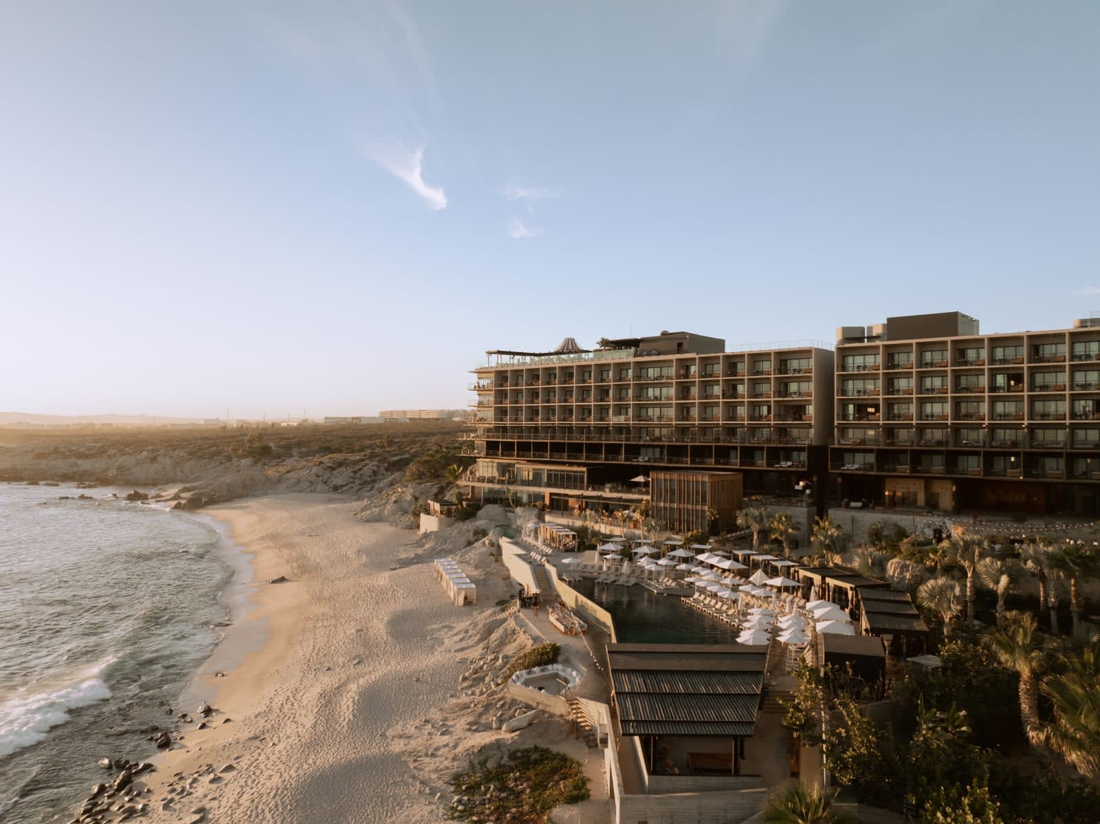 Clifftop beachfront resort at golden hour, with a multi-storey modern hotel overlooking a sandy cove, tiered pools and sun loungers below, and the ocean stretching along the rugged coastline to the left.