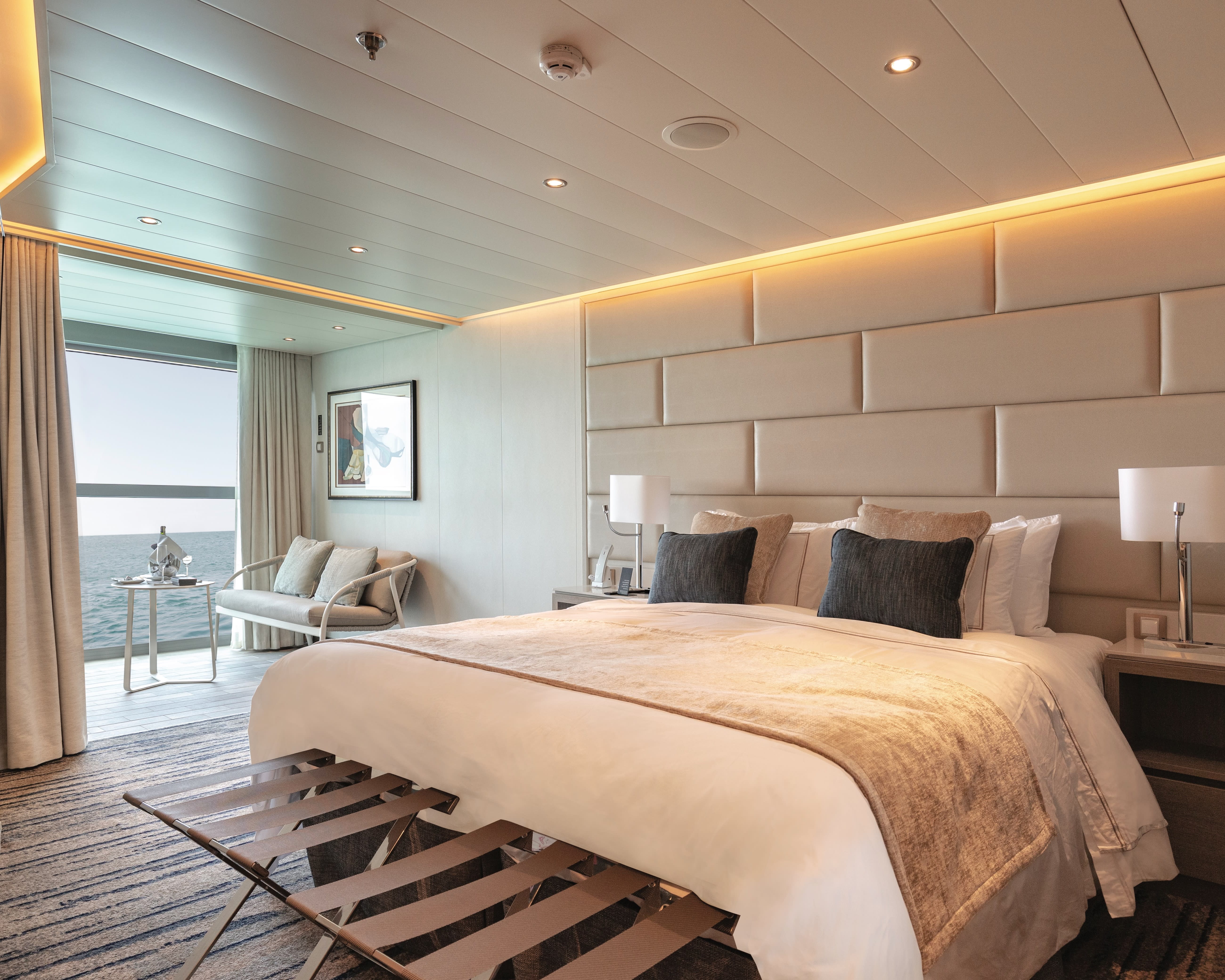 a contemporary bedroom with cream padded wall, white linen bed with dark grey accent pillows, and a loveseat next to a window with ocean water outside