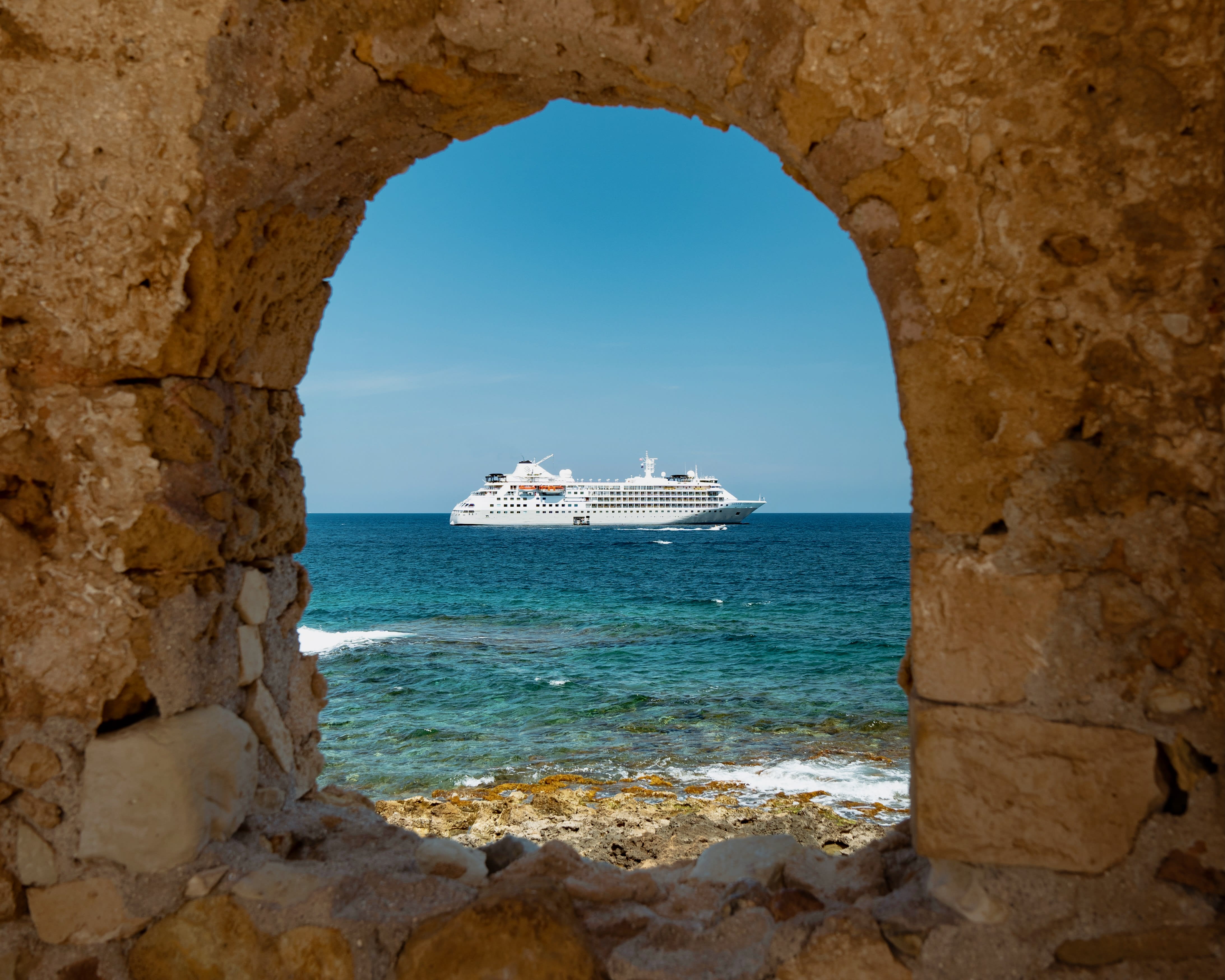 A cruise ship offshore in turquoise waters seen through an arched window in an ancient stone fort