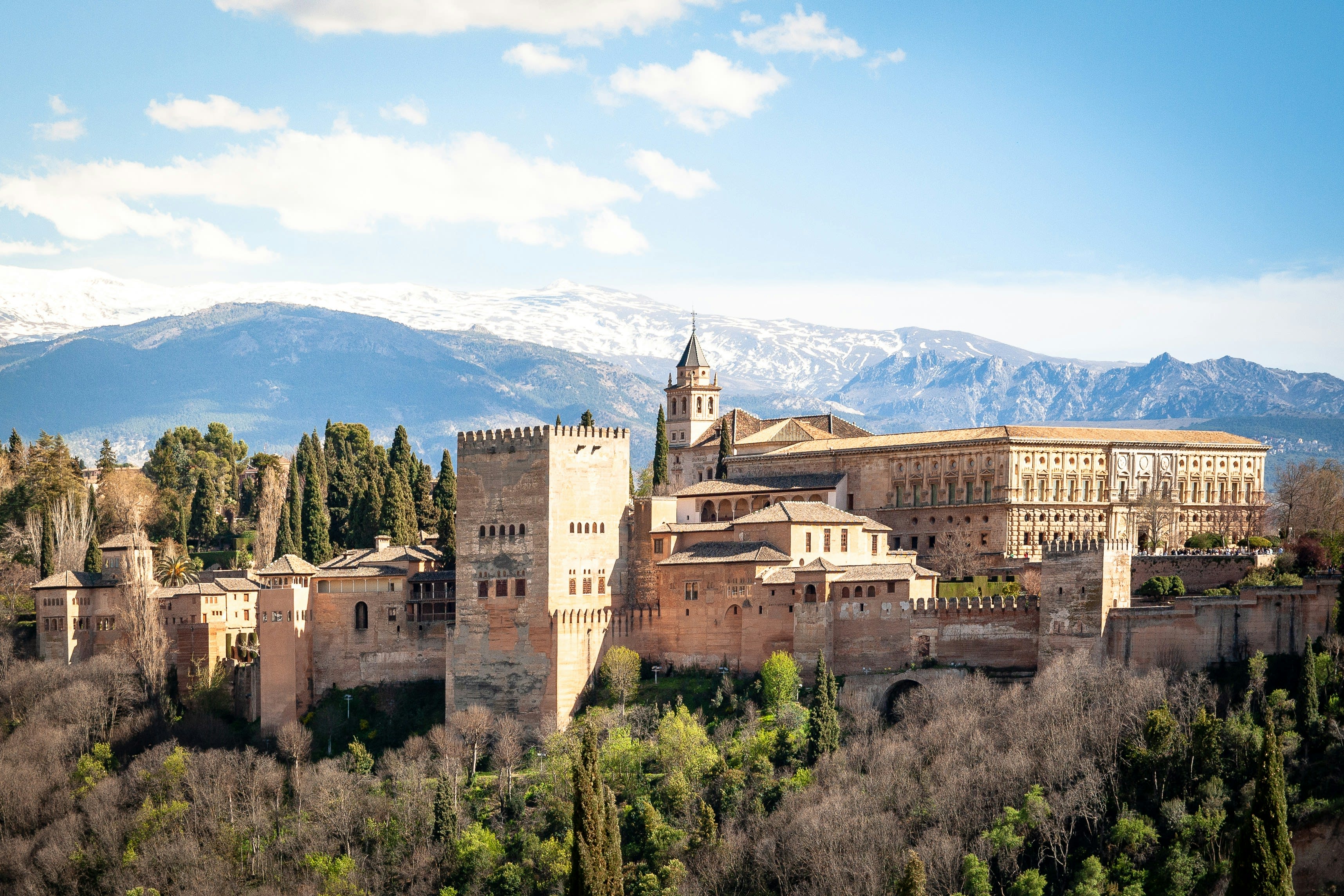 a cream colored fortress with trees in the foreground and snow capped mountains in the distance
