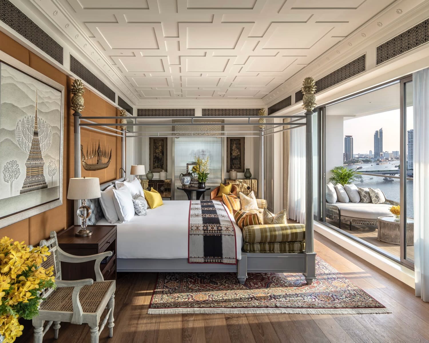 Thai hotel room with four poster bed, artwork on the wall, and wood floors overlooking the river