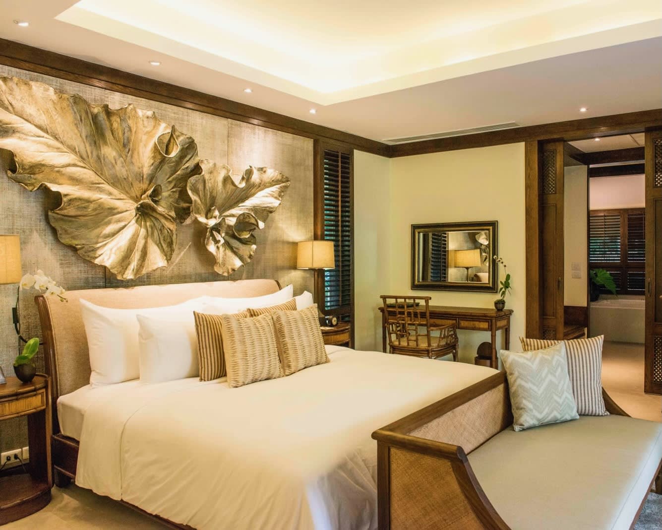 bench at the foot of a white linen covered bed and leaf decorated wall art above the bed