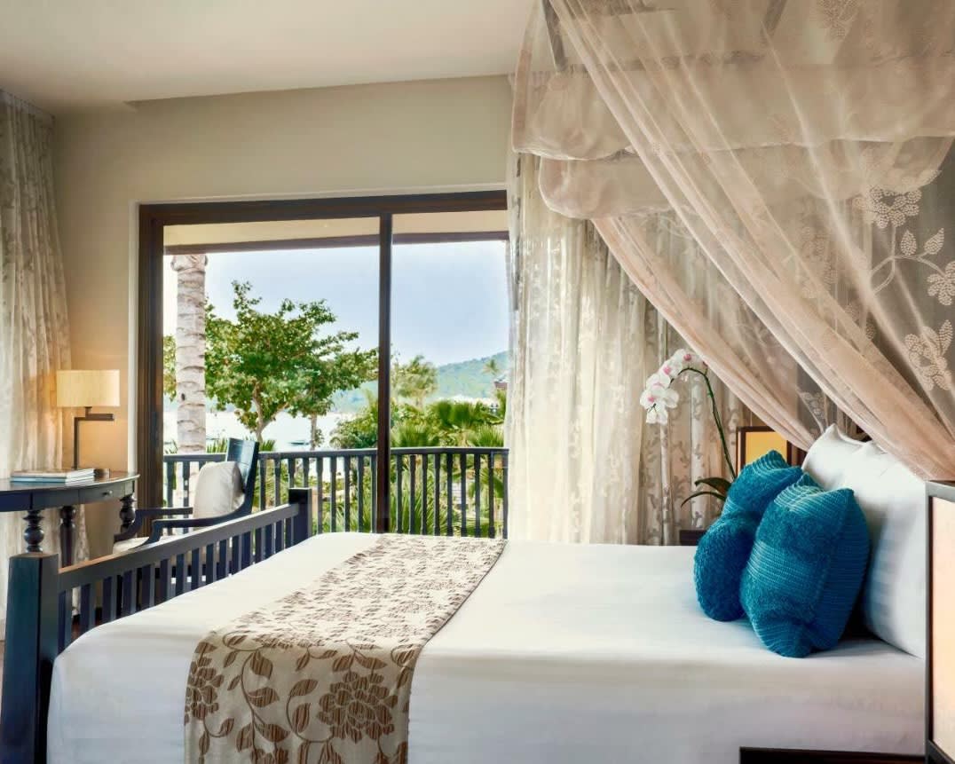 white bed with blue pillows and curtain over the head of the bed, next to a sliding glass door with trees outside