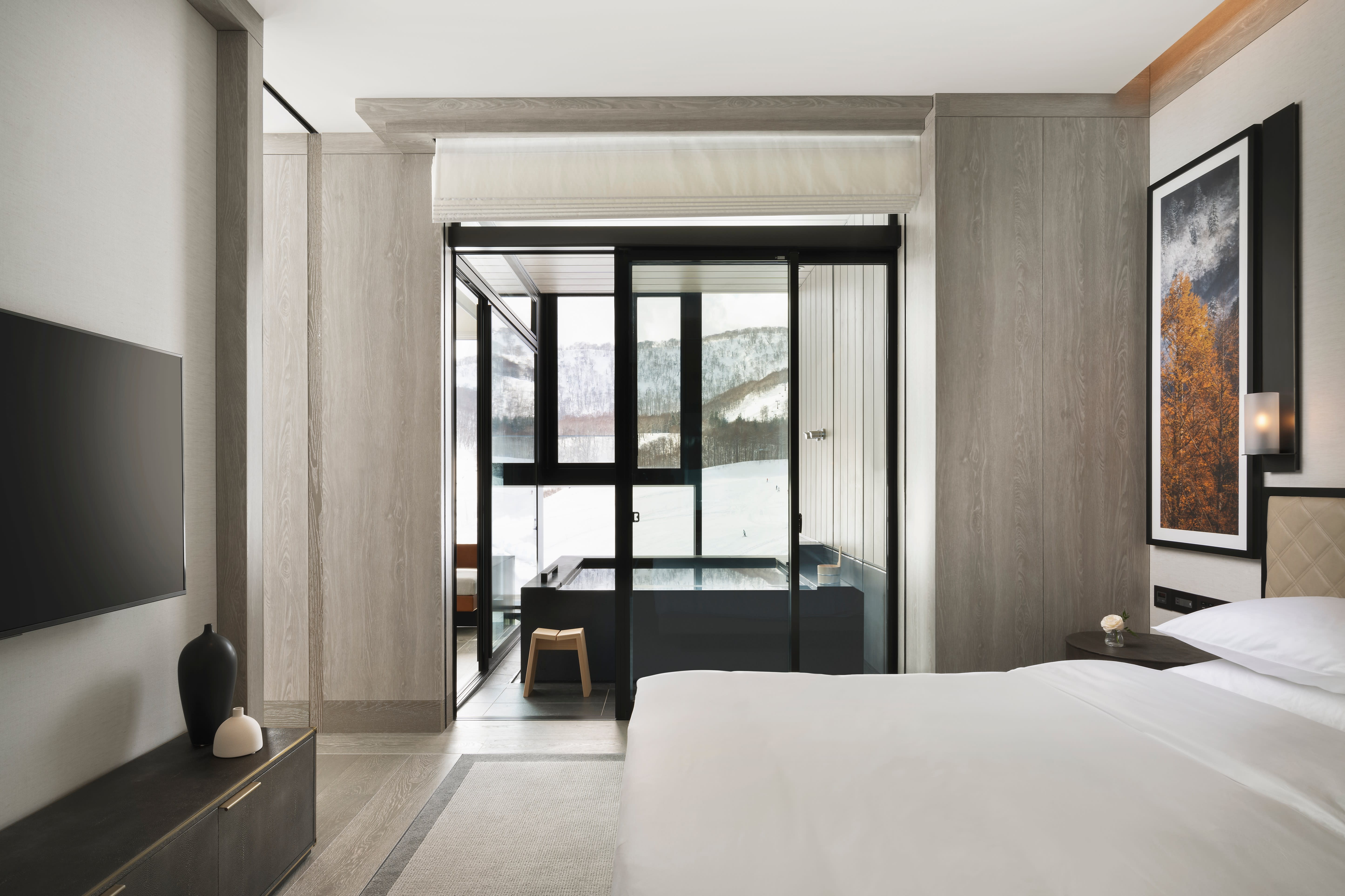 Sleek, minimalist hotel bedroom with a king bed, warm wood paneling, and a glass-enclosed soaking tub overlooking snow-covered mountains.