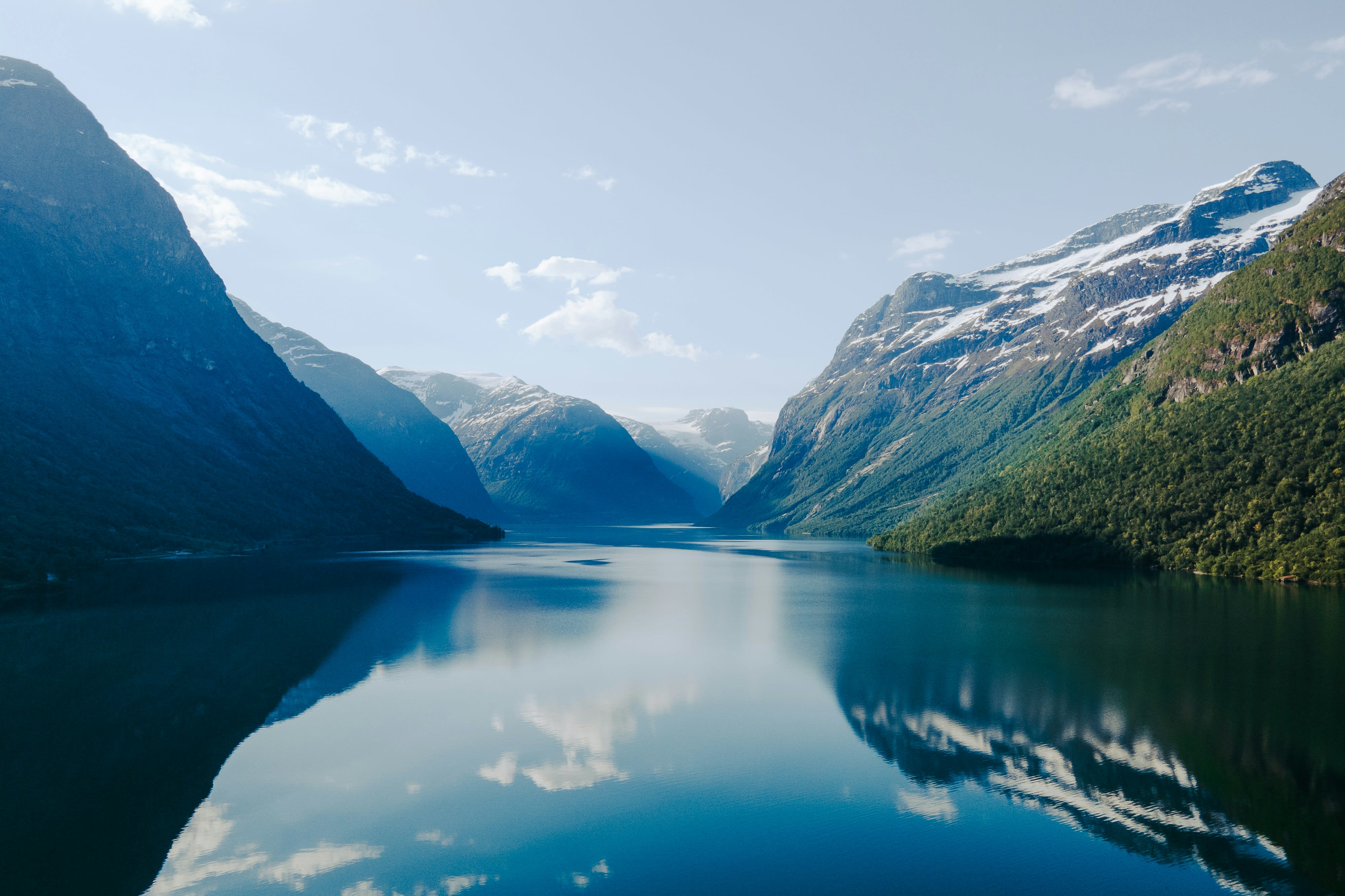 Serene fjord landscape with steep, forested mountains and snow-dusted peaks reflected in a calm, deep-blue lake under a clear sky.