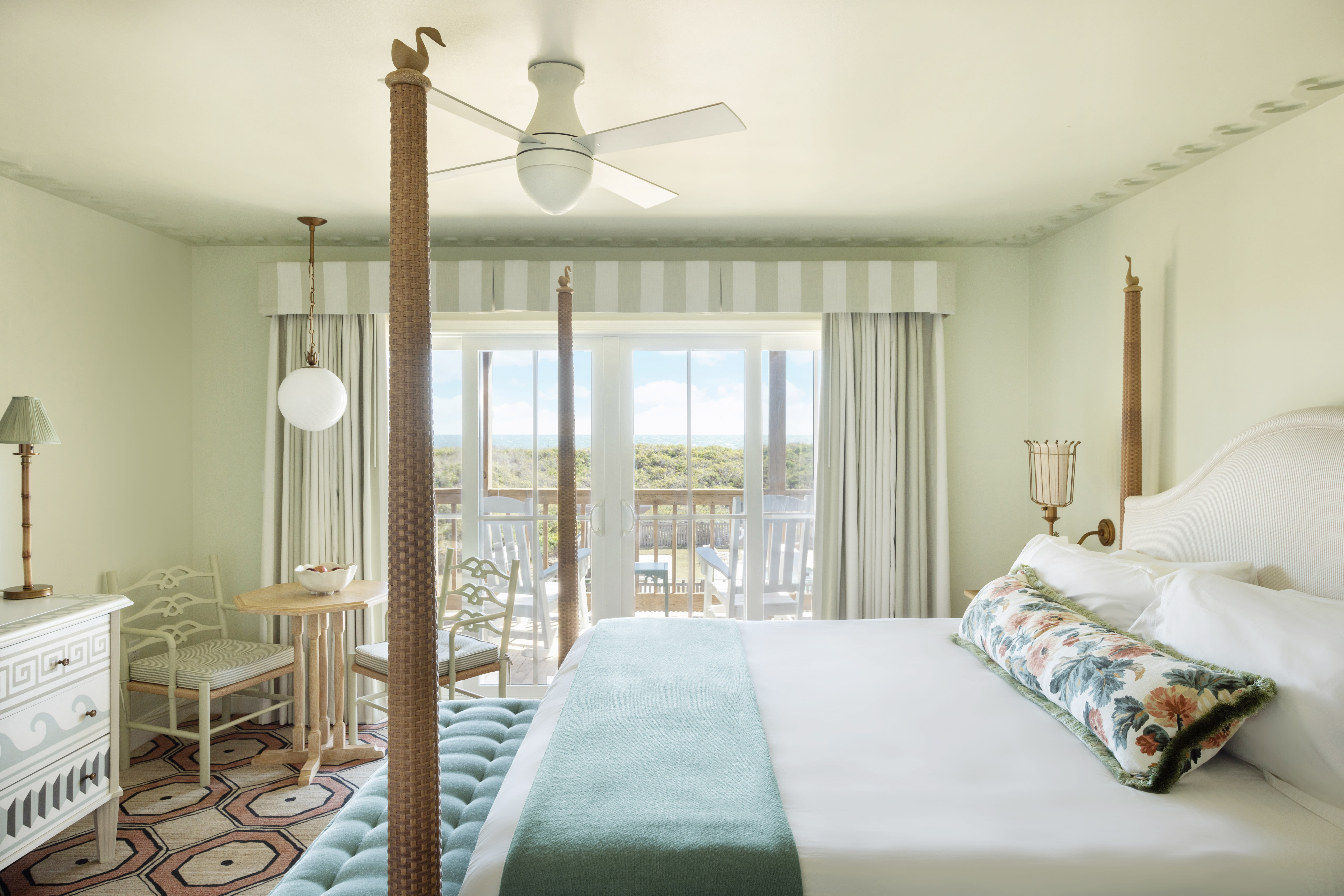 Bright coastal-style bedroom with a four-poster bed, soft green accents, patterned rug, and French doors opening onto a balcony with treetop and ocean views.