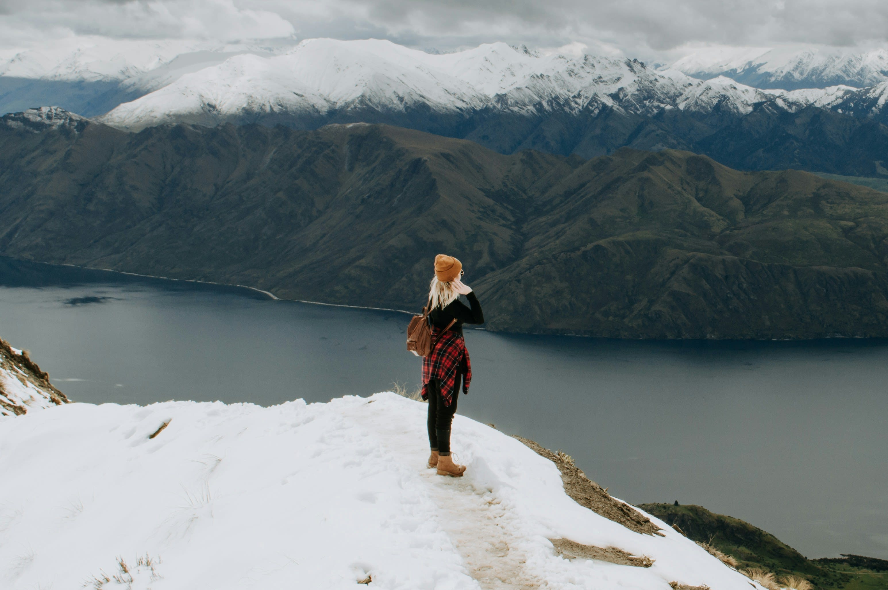 Person standing on a snowy cliff edge, looking out over a deep blue lake and dramatic snow-capped mountains under a cloudy sky.