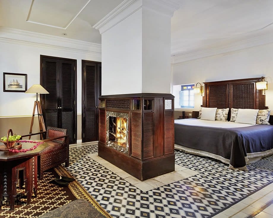 wood walled fireplace in the middle of a hotel room with wood chair and table off to the left and blue covered bed with wood headboard off to the right and blue and white tile floors