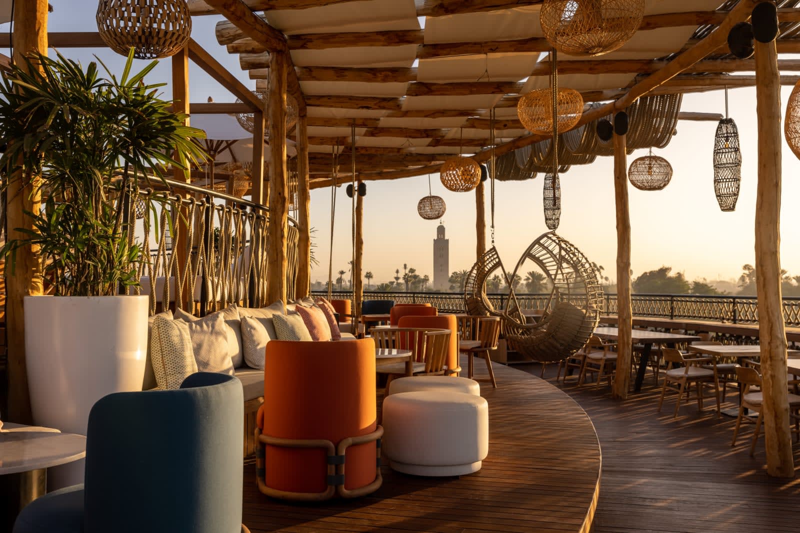 Rooftop terrace at Nobu Marrakech with woven lanterns, timber pergola beams, low lounge seating, and hanging rattan chairs, set against golden-hour views of the Medina and Koutoubia Mosque in the distance.