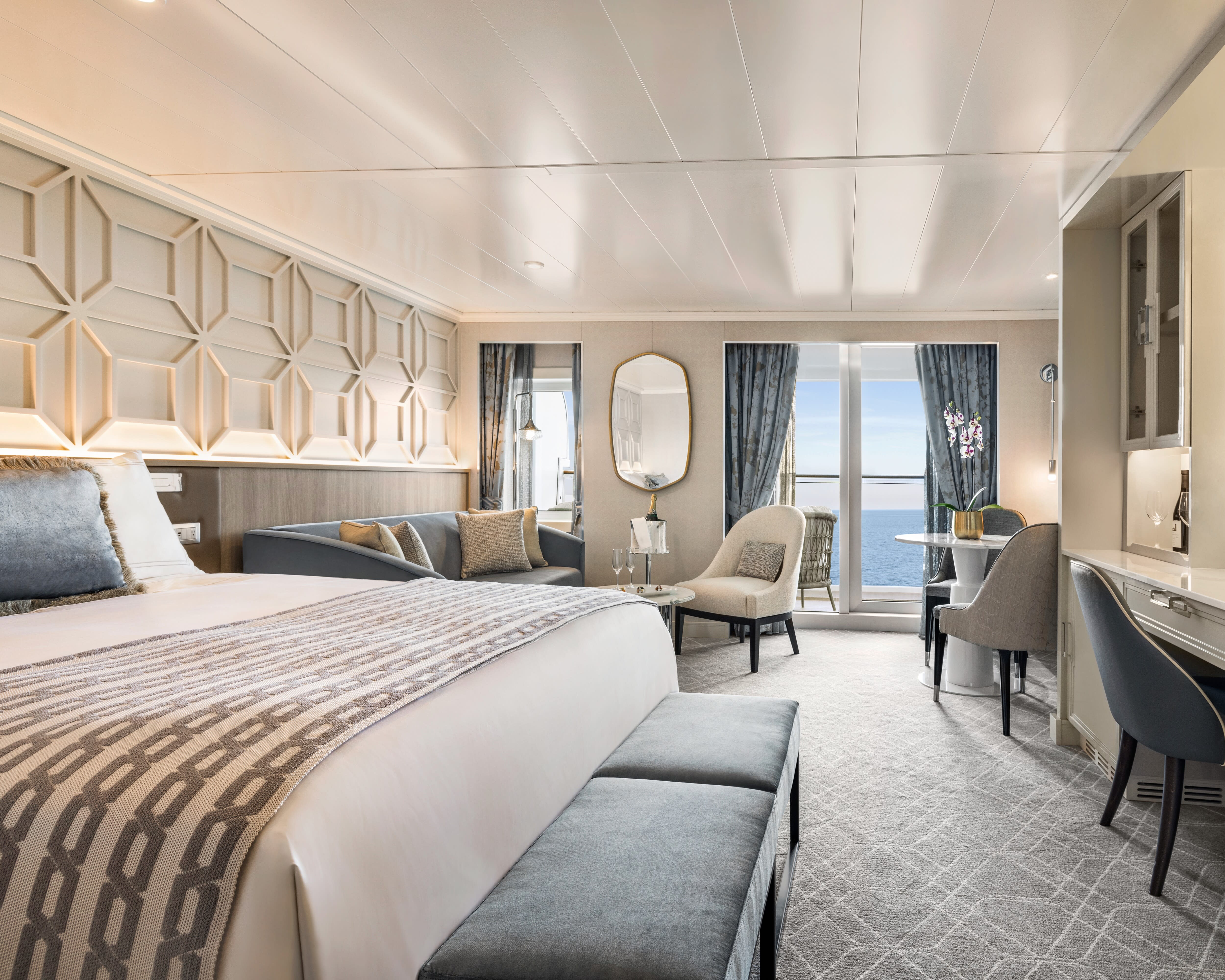 modern cruise suite with bed off to the left, sofa and chair next to a window, and table with chairs next to a glass balcony door