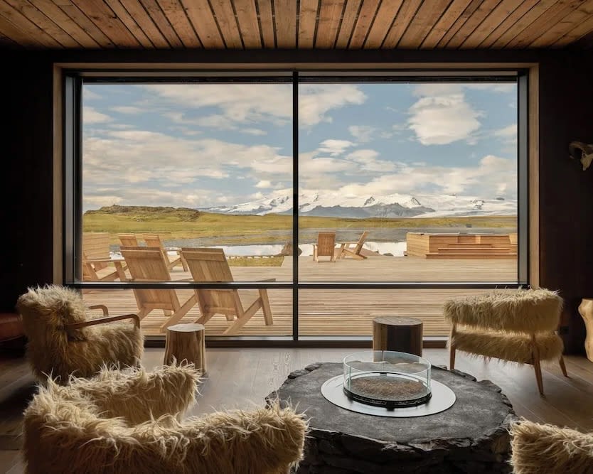 lounge with cream faux fur chairs and fire pit next to large window with outdoor deck and chairs overlooking a green landscape