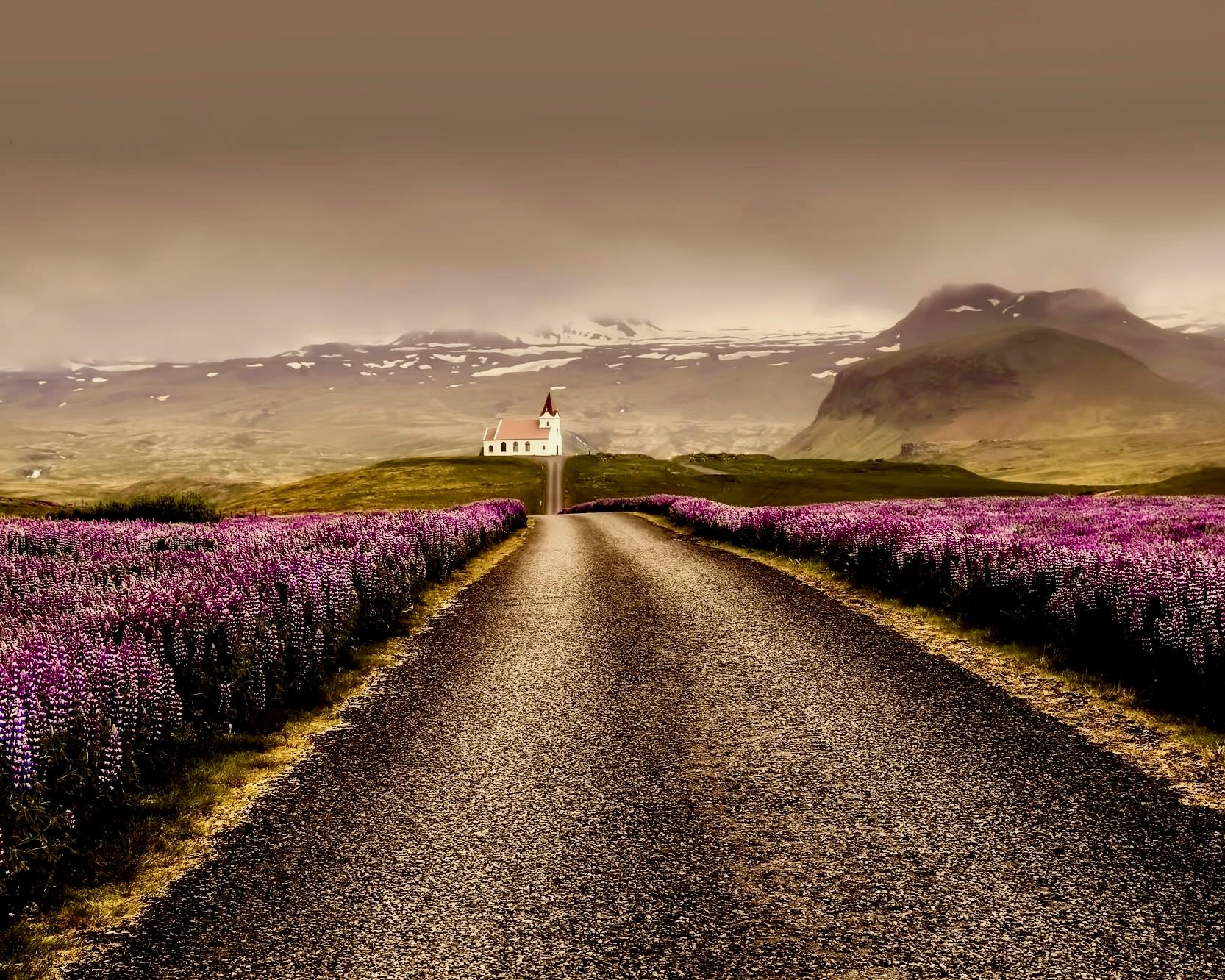 black road lined with purple flowers leading to a distant white church and green mountains with cloudy skies