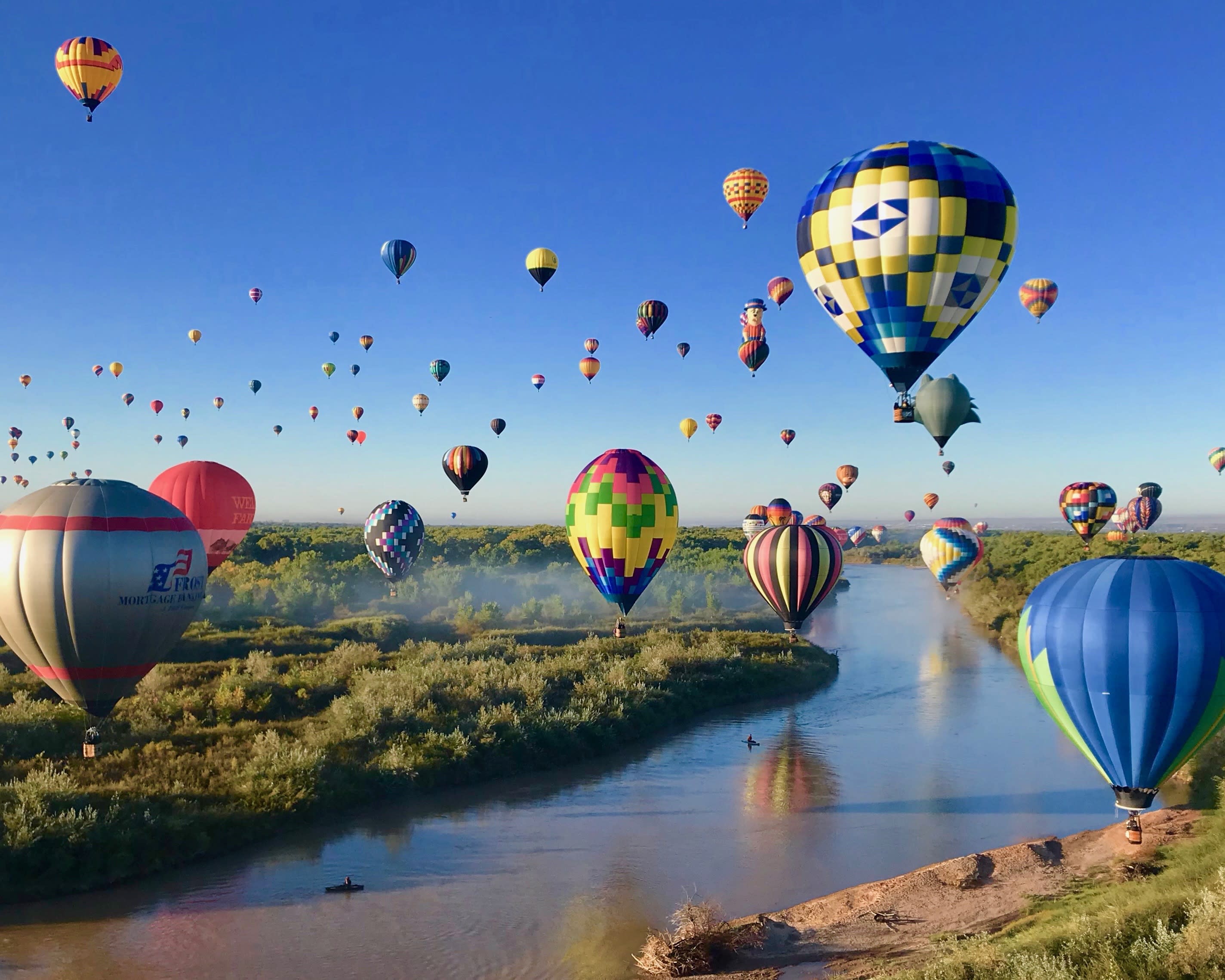 colorful hot air balloons taking to the skies above a river with green grass on either side