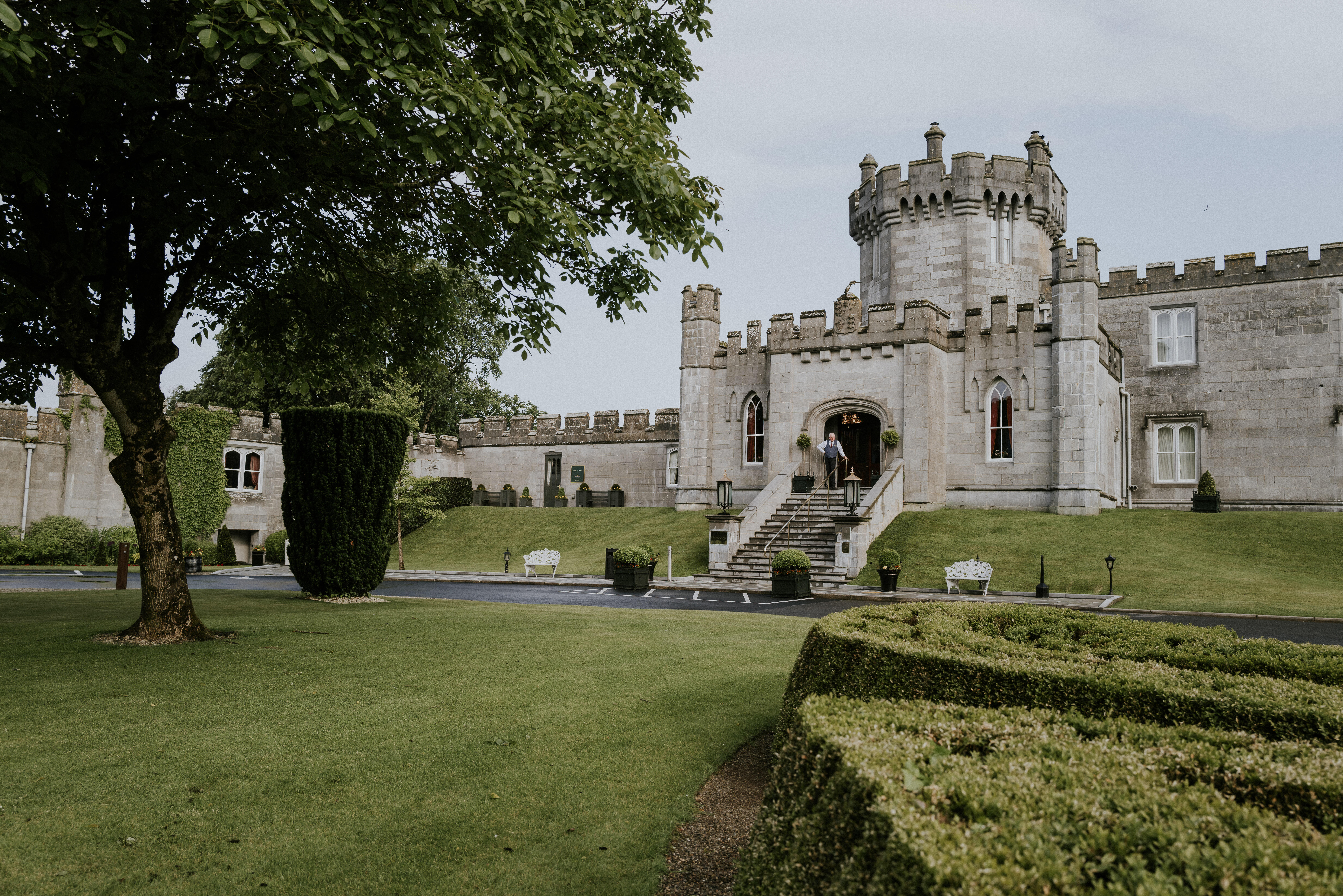 grand castle with multiple towers and a central entrance, surrounded by well-maintained lawns and hedges. The castle features stone walls, arched windows, and a staircase leading to the main entrance.