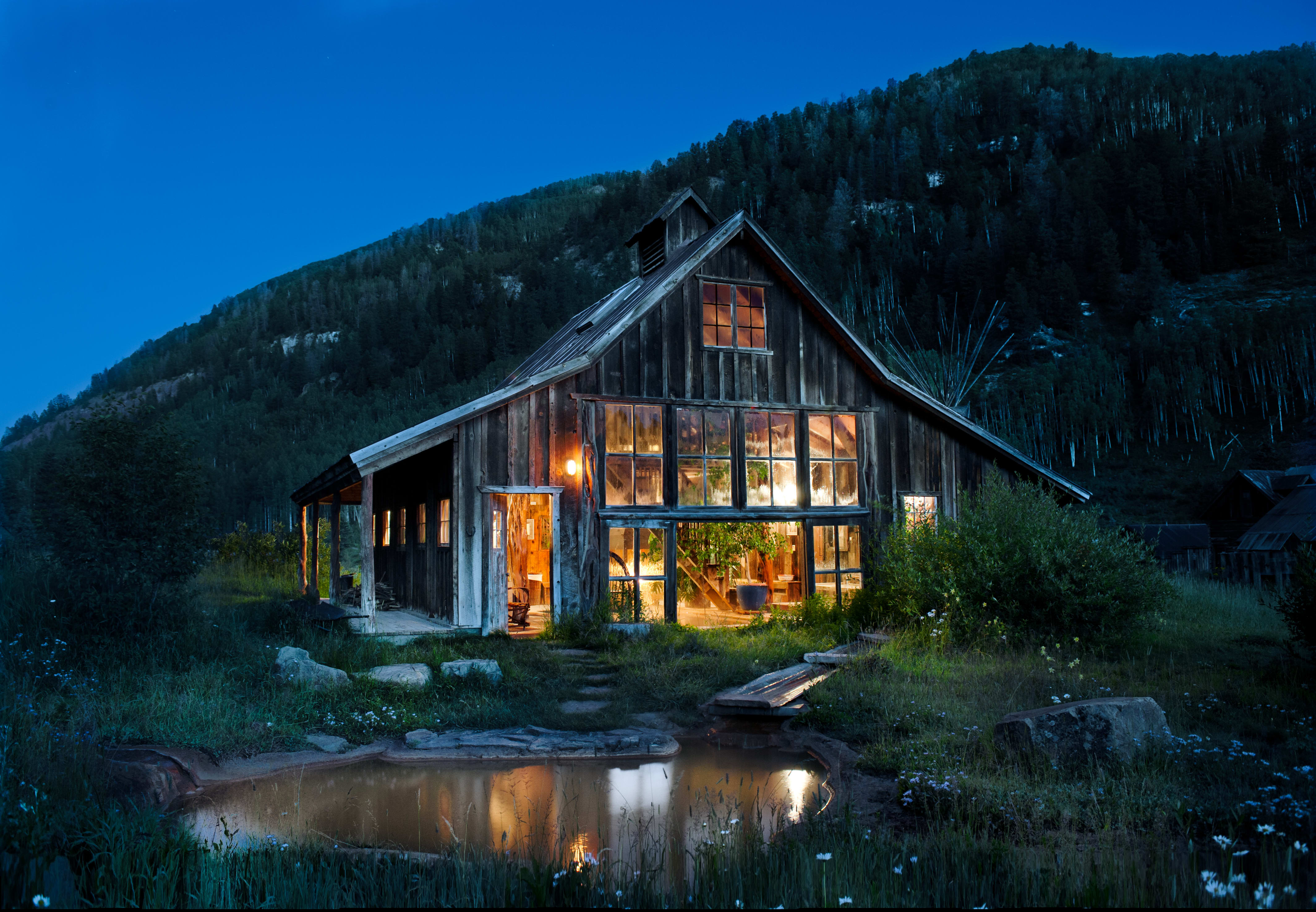 A rustic wooden cabin nestled in a grassy field at dusk. The cabin features large glass windows and doors that allow the warm interior light to glow invitingly against the darkening sky.