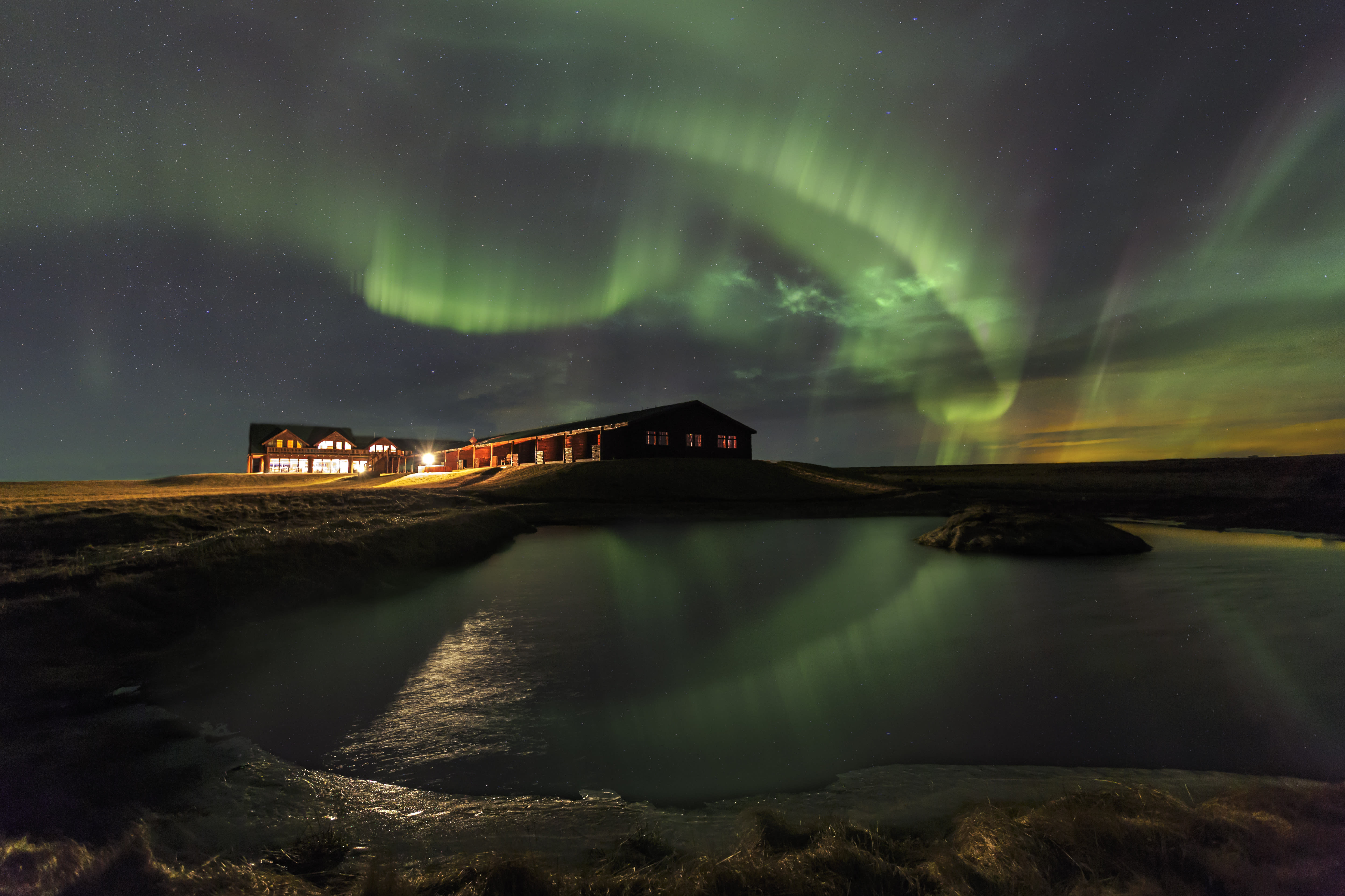 A serene night scene with a building illuminated by warm lights, situated near a calm body of water. The sky above is filled with vibrant green and yellow hues of the Northern Lights