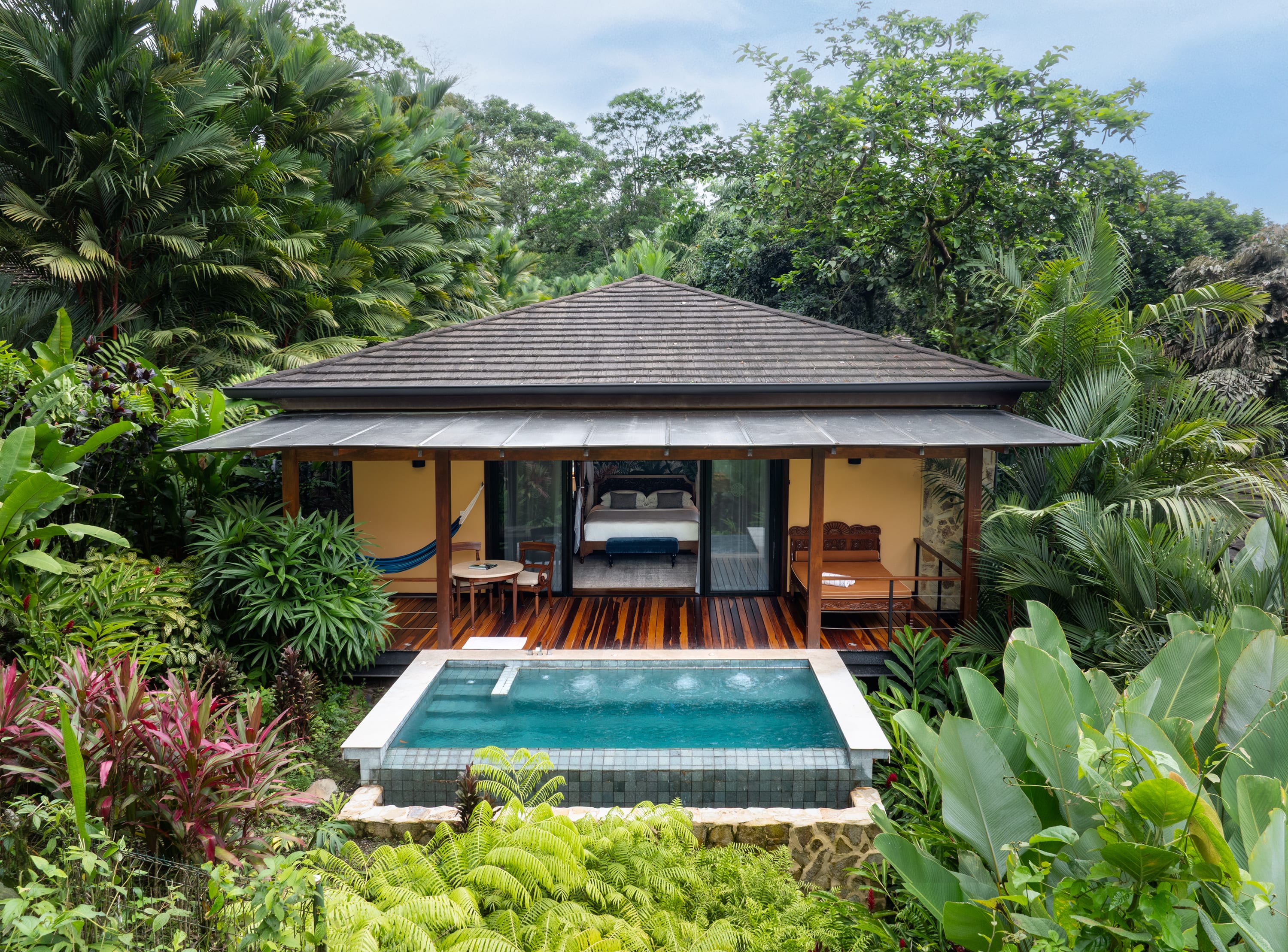A secluded bungalow nestled within a lush tropical garden. The bungalow features a thatched roof and wooden walls. In front of the bungalow, there is a small, inviting pool with clear blue water. 