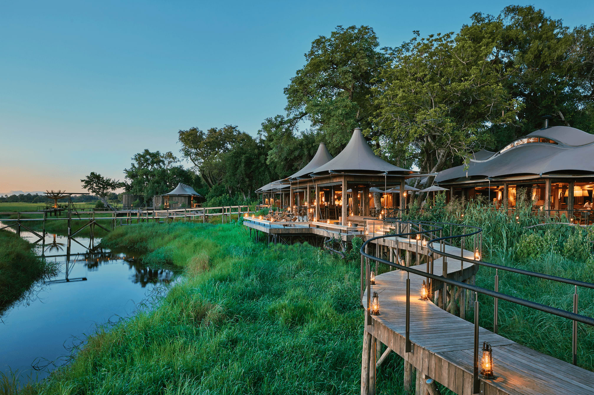 several tented pavilions with conical roofs, arranged in a row along a wooden boardwalk that winds through the greenery. In the foreground, there is a tranquil body of water reflecting the surrounding scenery. 
