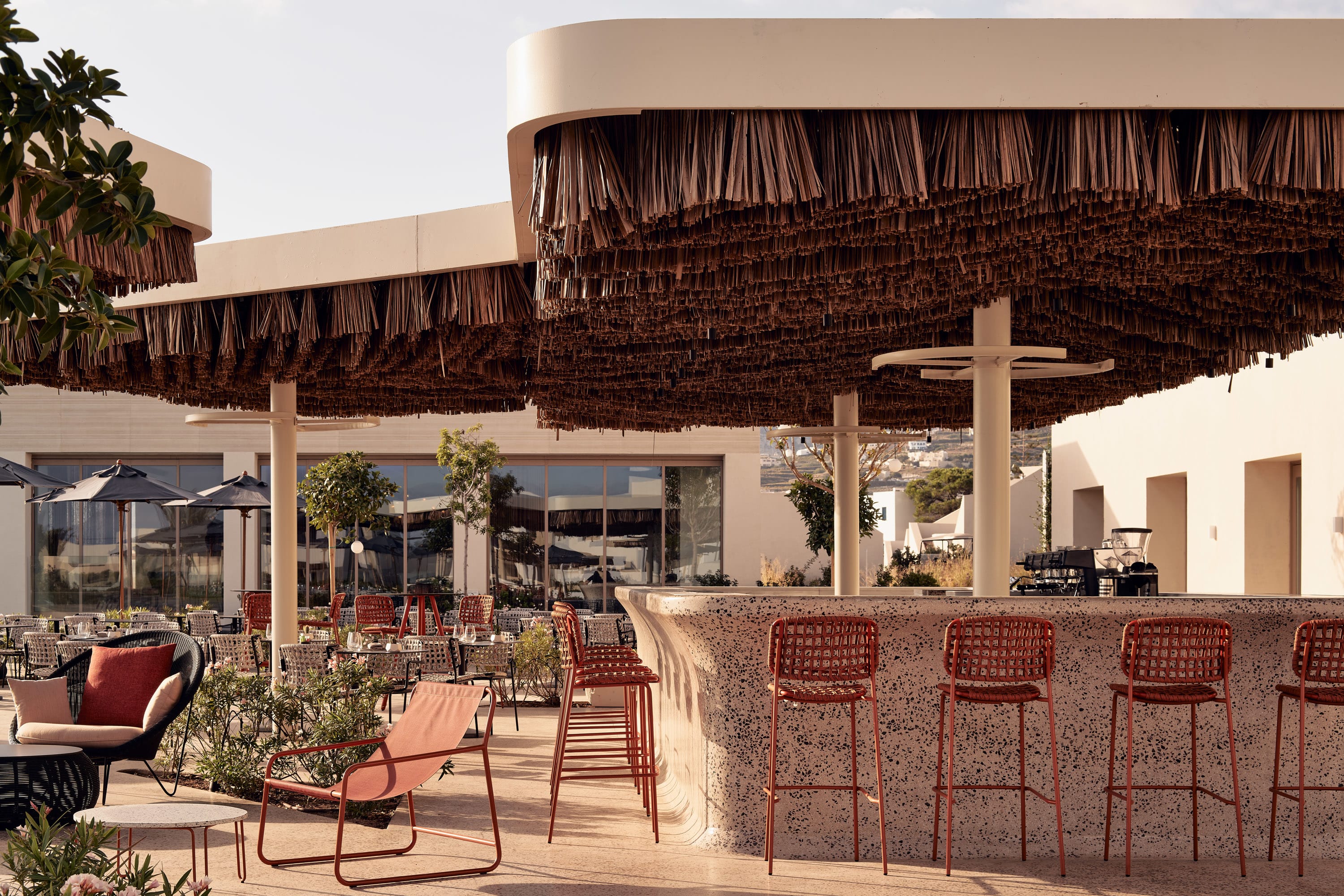 An open-air poolside bar with a curved terrazzo counter and woven red bar stools sits beneath a dramatic thatched canopy, surrounded by low lounge seating and sunlit whitewashed buildings in the background.
