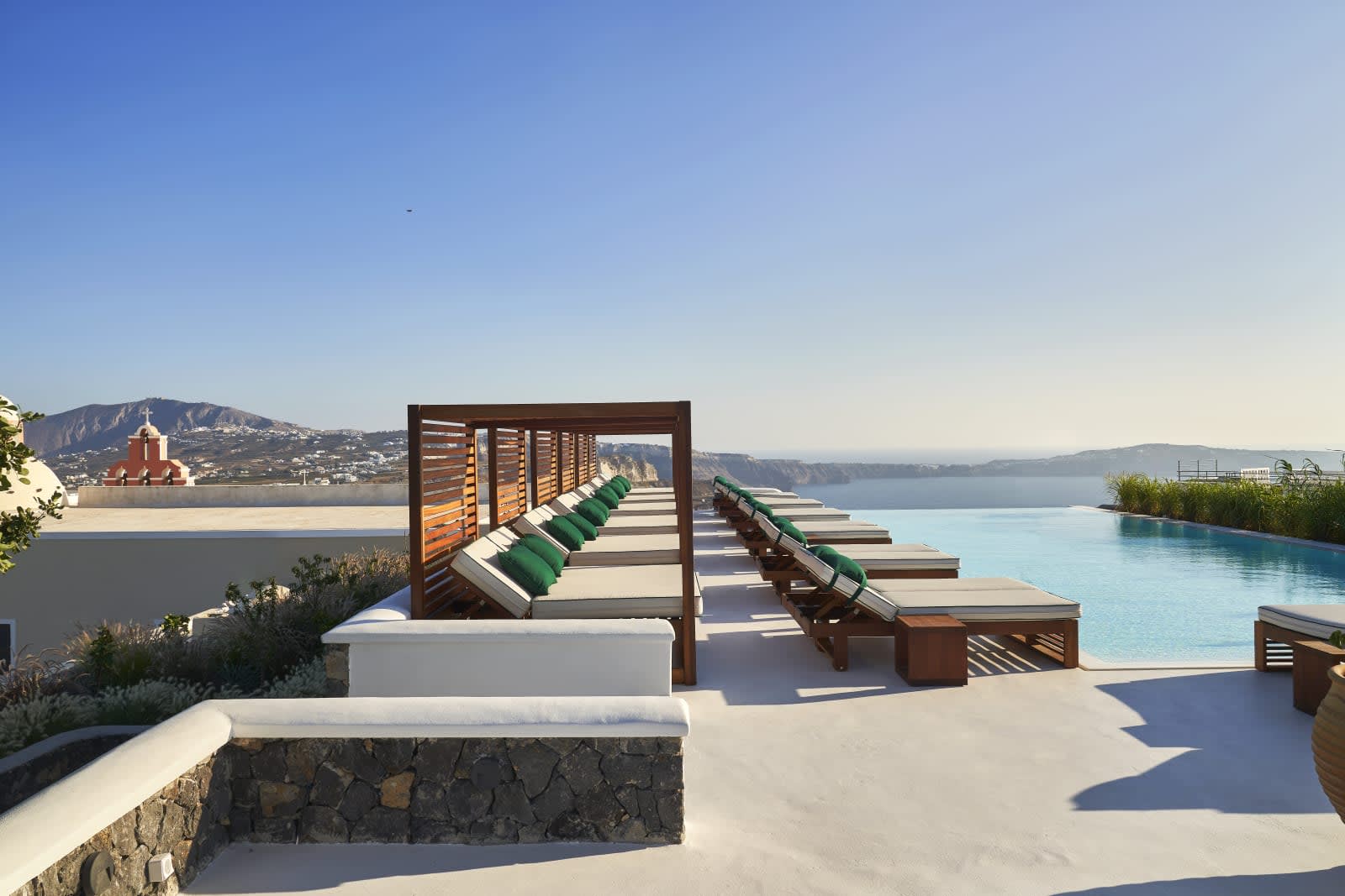 A row of cushioned wooden loungers with green pillows lines an infinity pool overlooking the sea, with whitewashed terraces and a distant cliffside village under a clear blue sky.