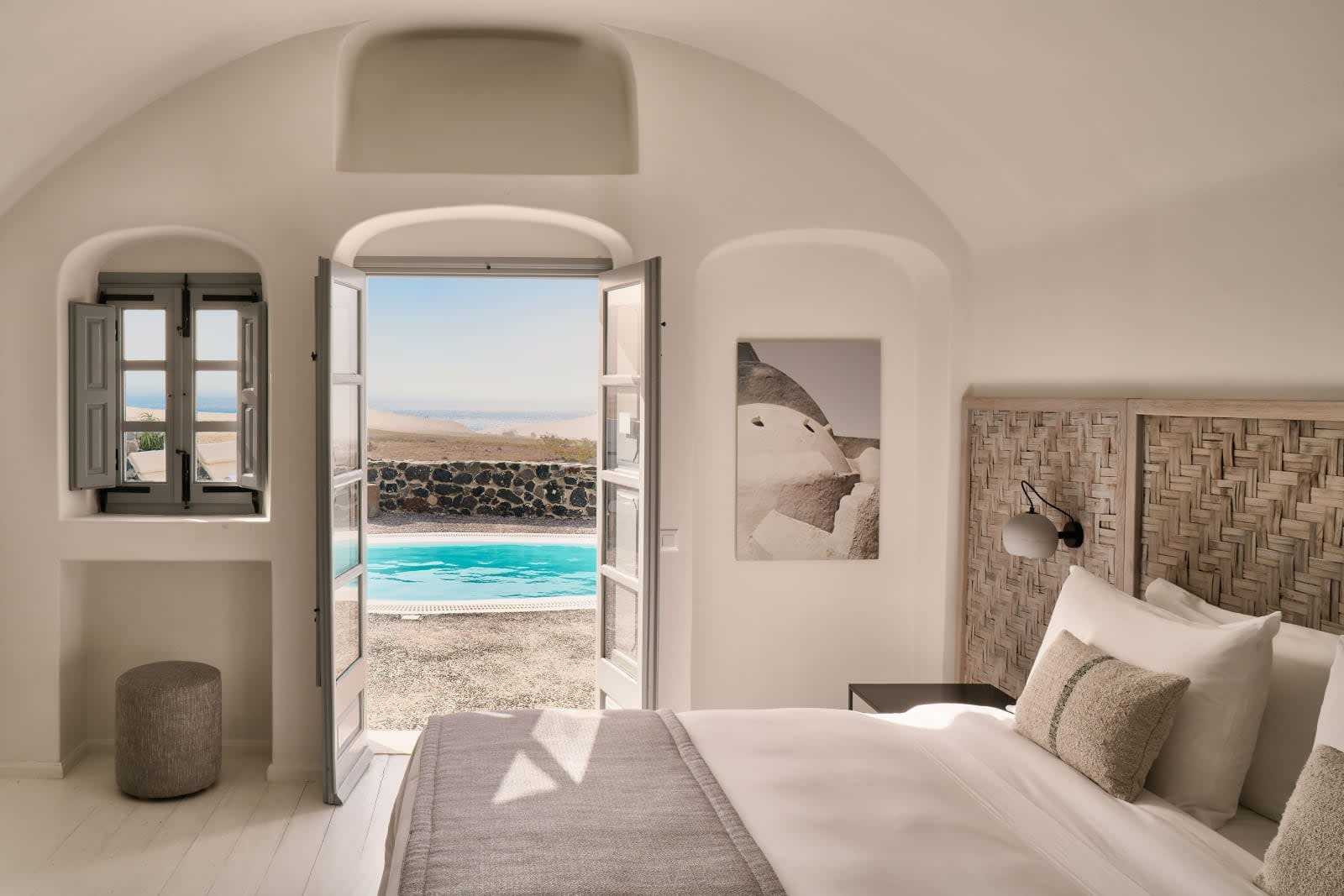 Minimalist white bedroom with woven headboard and neutral linens, French doors opening to a small private pool and sea views beyond a stone wall under bright Mediterranean light.