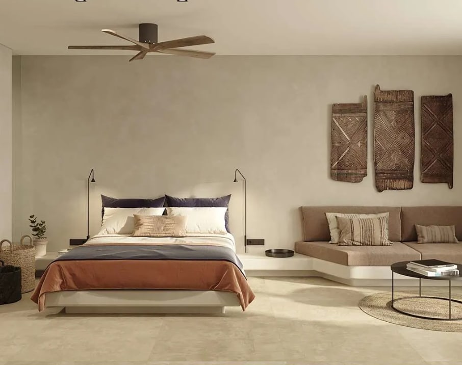 Contemporary neutral-toned bedroom with low platform bed dressed in layered earth-tone linens, minimalist wall art, built-in sofa seating and soft ambient lighting beneath a ceiling fan.