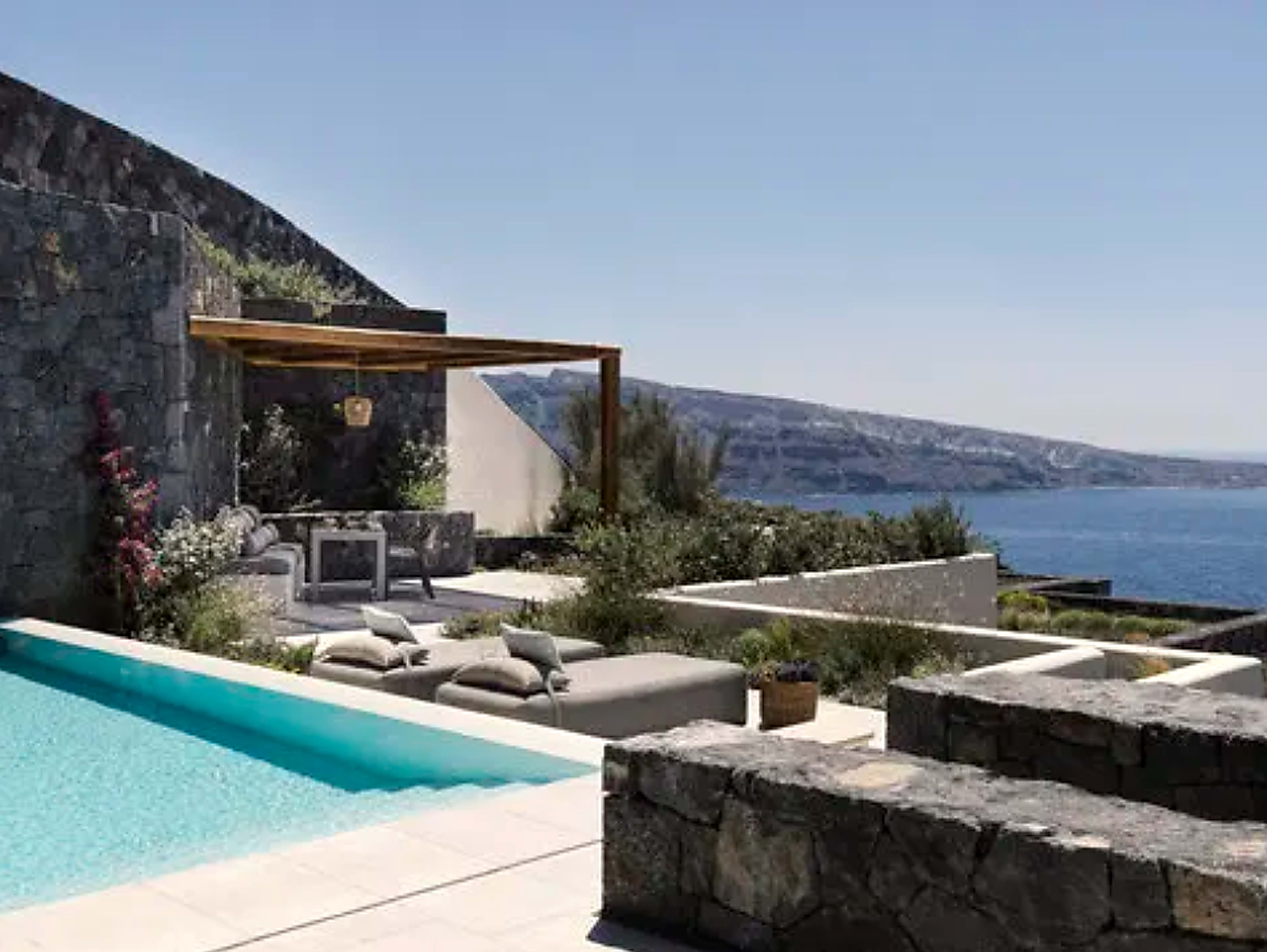 Private cliffside terrace with infinity pool, stone walls and shaded lounge area overlooking the sea and rugged coastline under clear blue skies.