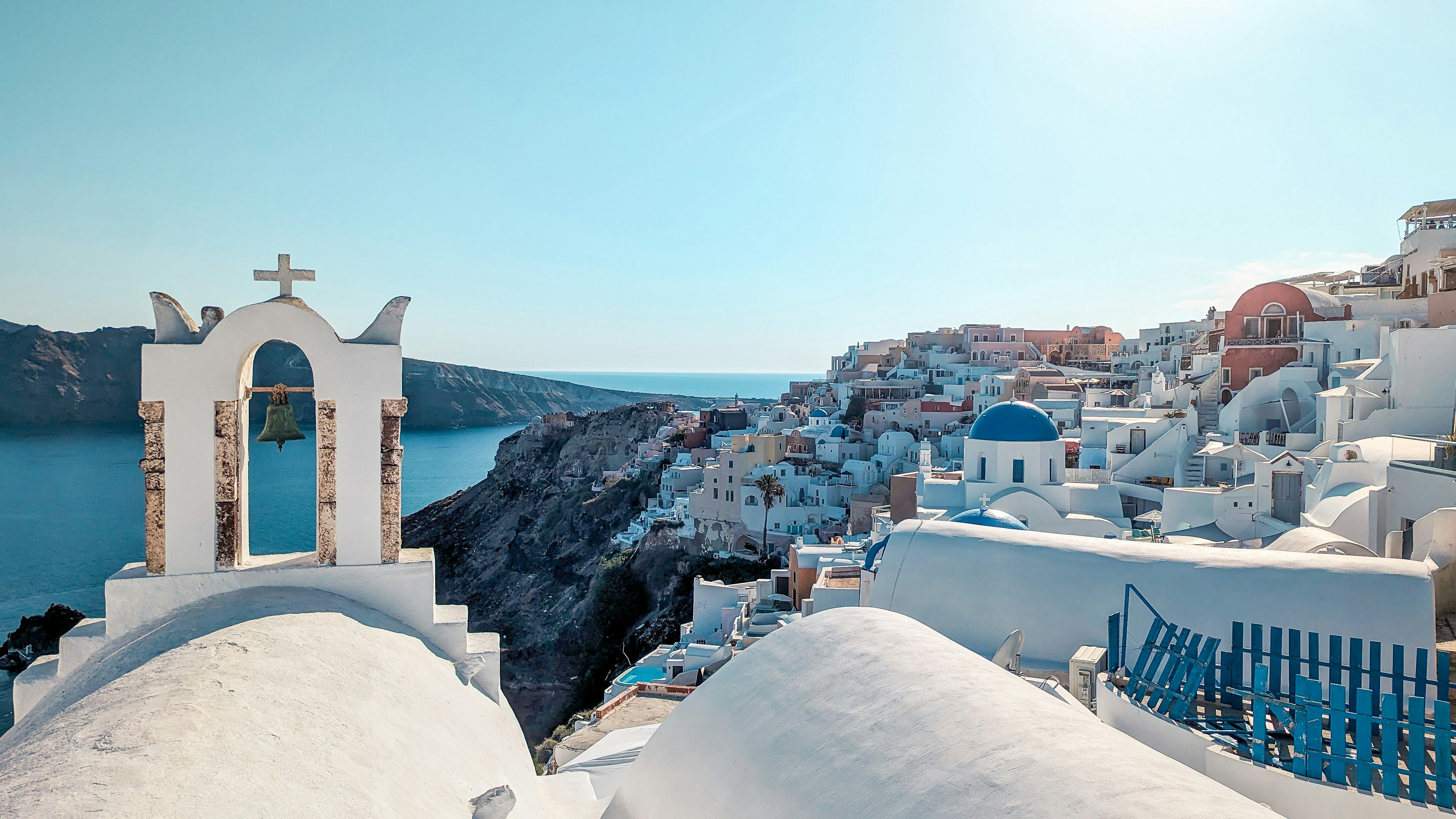 Whitewashed church bell tower and domed rooftops overlooking Santorini’s cliffside village and the deep blue Aegean Sea under a clear sky.