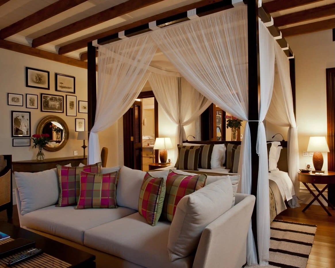 dark wood four poster bed with shear white curtains, a white sofa at the foot of the bed, wood beam ceilings, and white walls