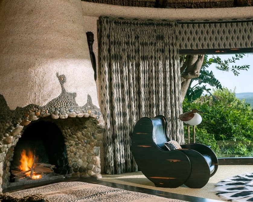 green chair looking out floor-to-ceiling windows at green trees, next to a drawn curtain and stone fireplace