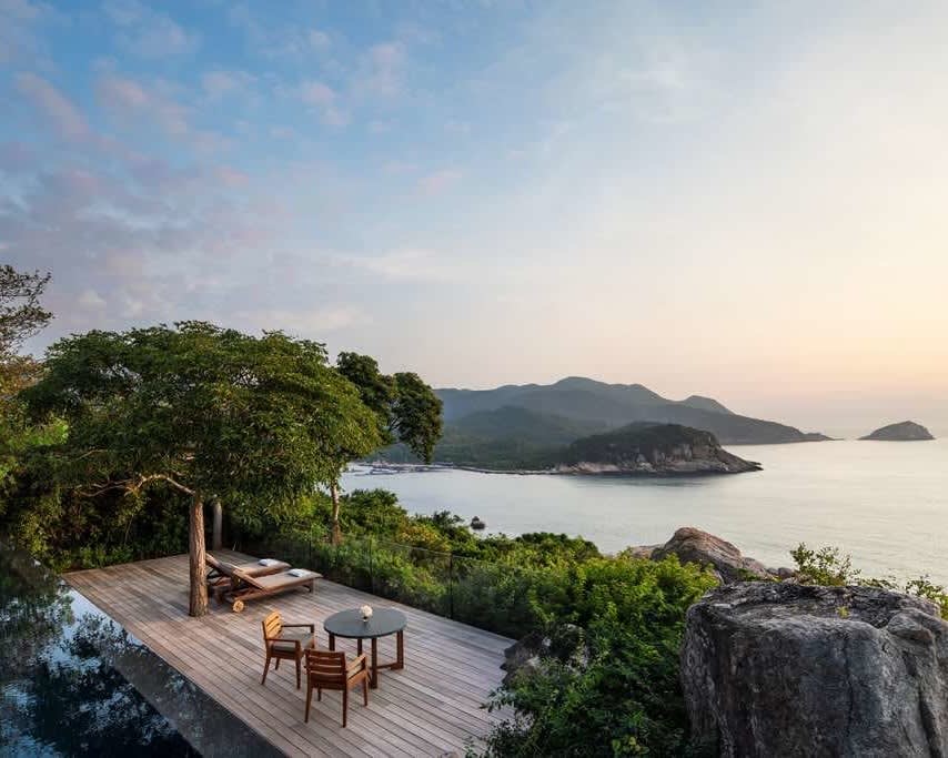 villa pool deck with chairs overlooking a tropical ocean landscape with trees, mountains, and the sun setting