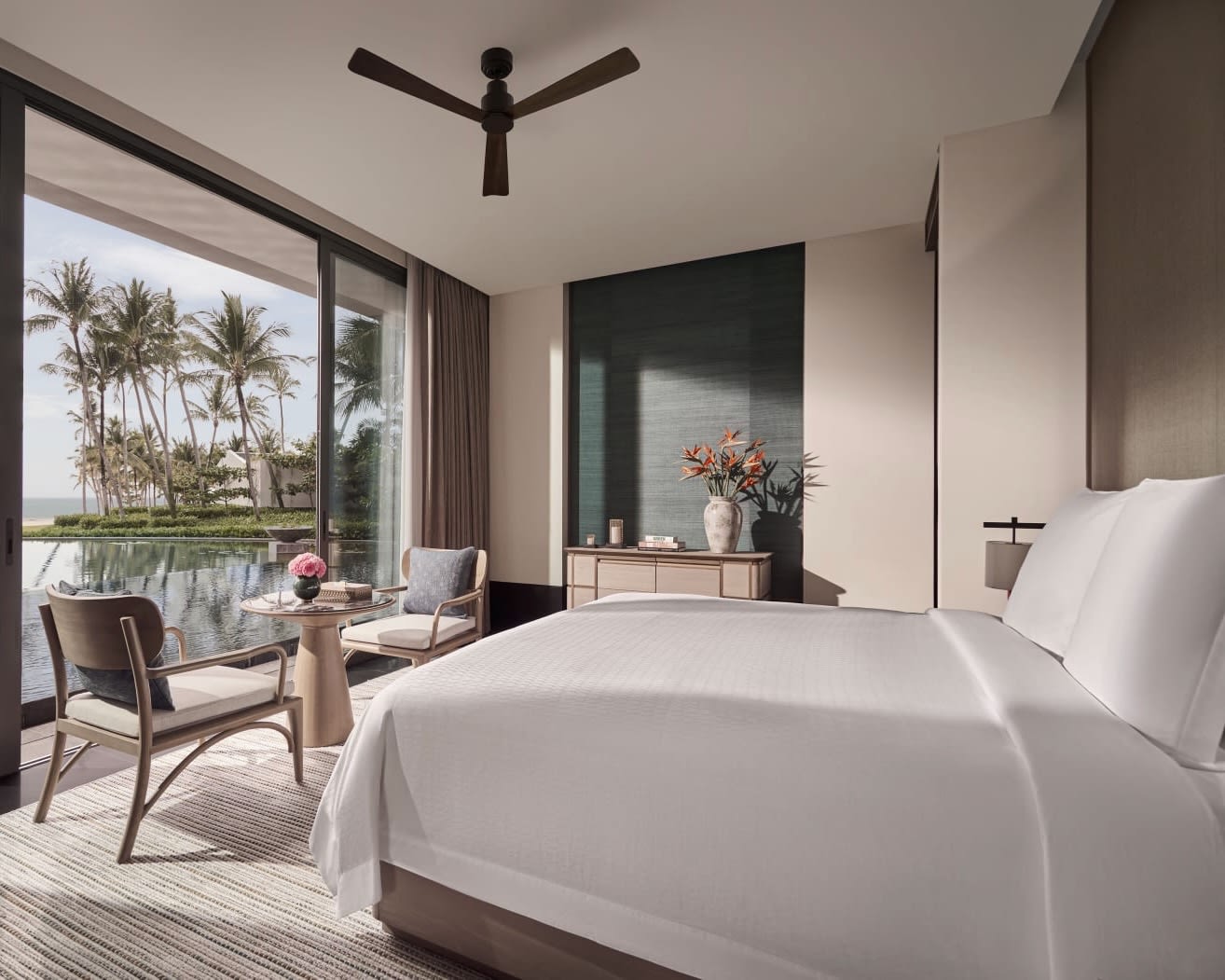 white bed facing a pair of chairs and table next to a window overlooking a pool with palm trees