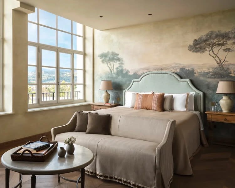 beige bed with green headboard and orange pillows in front of a landscape fresco wall