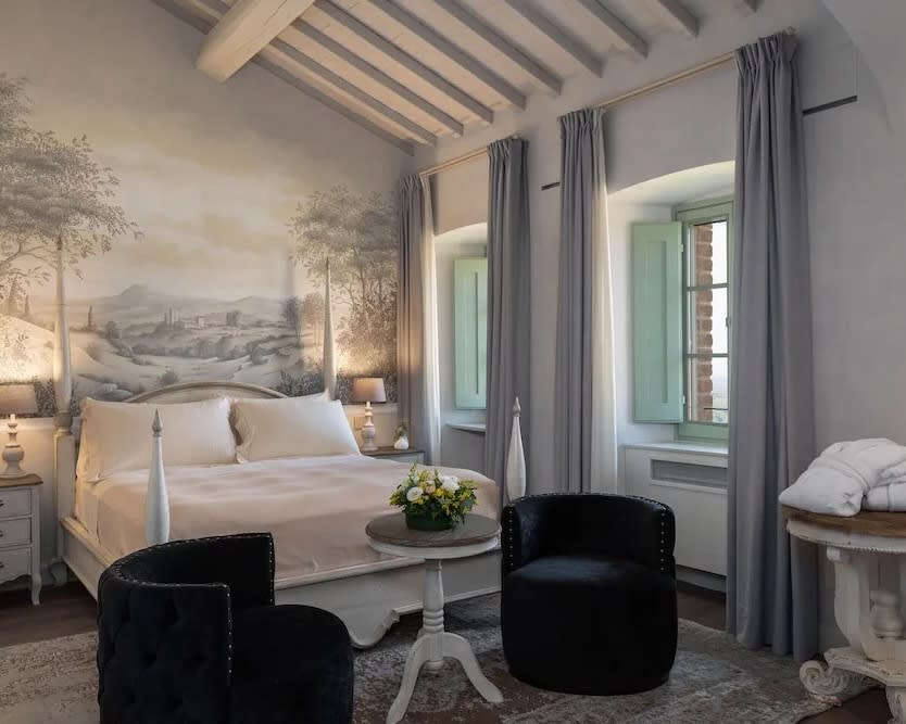 white four poster bed with two black chairs at the food of the bed, grey curtains, and wood beam ceiling with a fresco painted wall