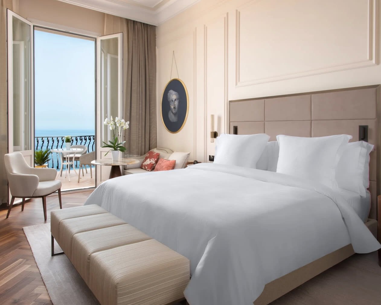 tan bed frame with white sheet covered bed next to table and chairs beside a balcony overlooking the ocean during day