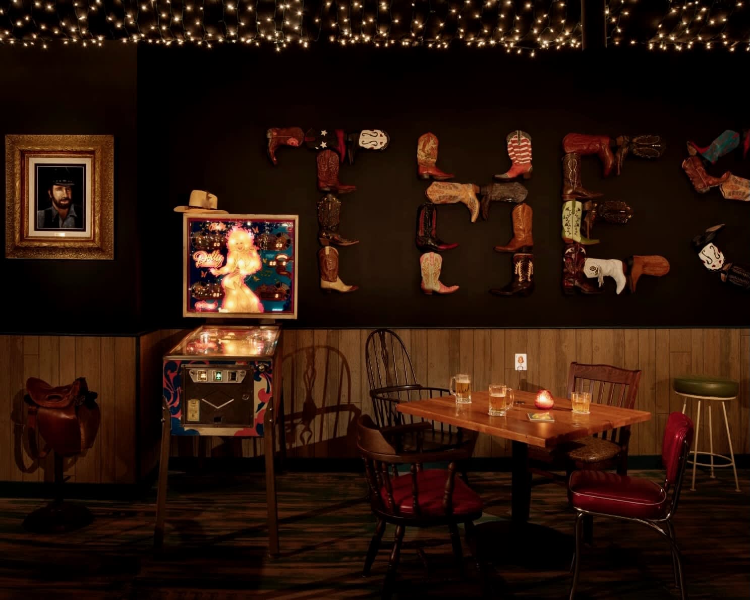 southern country bar with table and chairs, pinball machine, and cowboy boots as wall decor