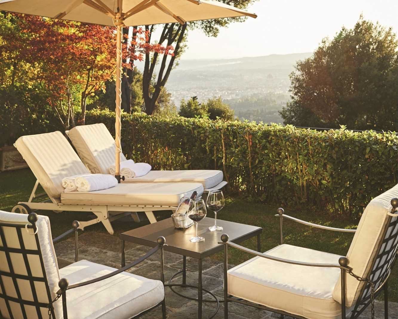 white cushion and black metal chairs, a black table with wine glasses, and two white lounge chairs under an umbrella overlooking the countryside