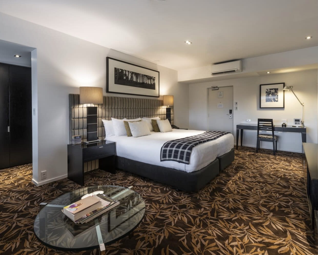 hotel suite with dark patterned carpet, glass table, and large grey bed and desk along far wall