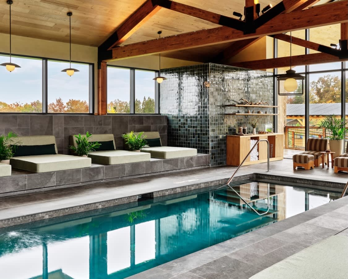 indoor spa pool with loungers lining one side, with wood beam ceiling and large windows