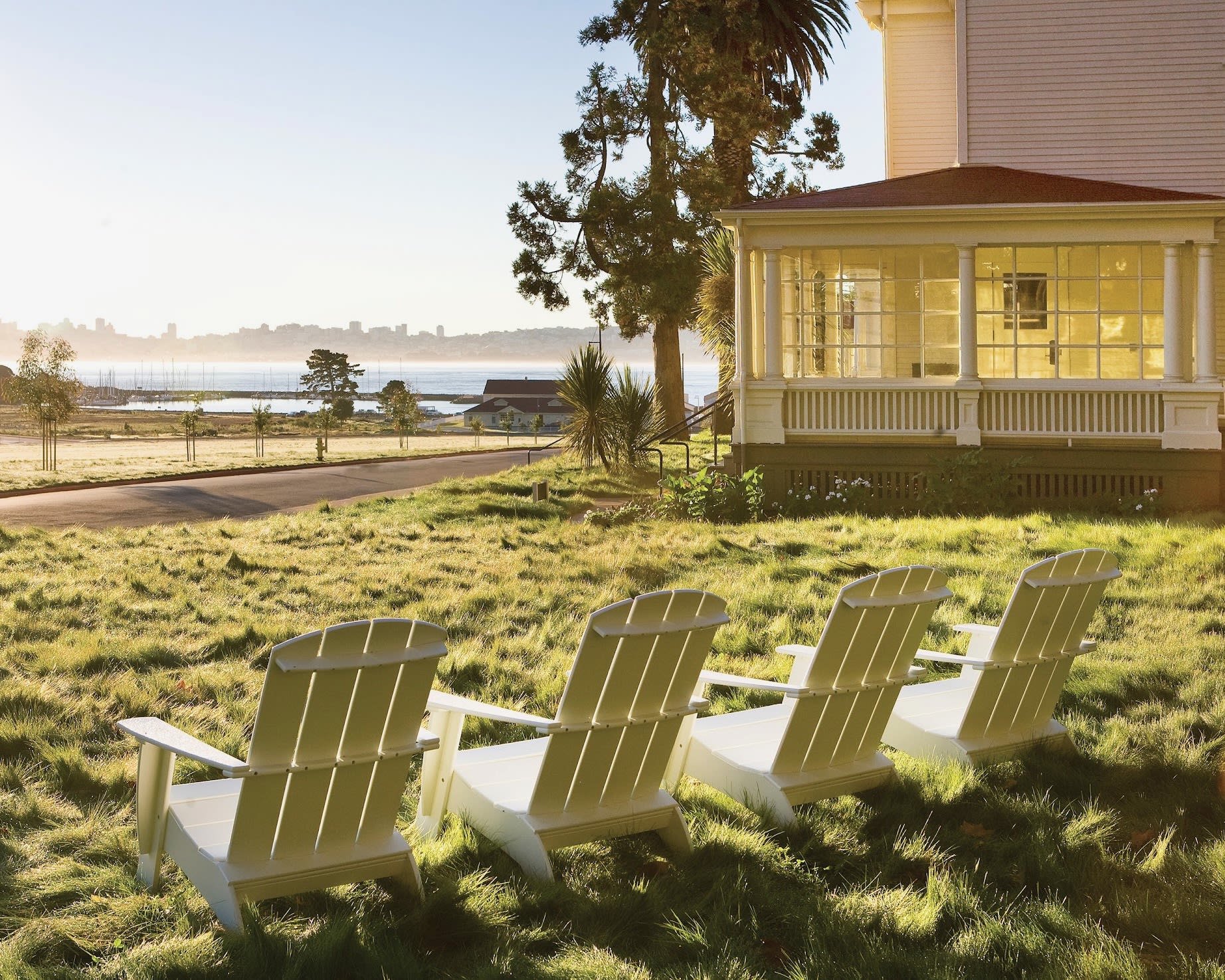 white adirondack chairs on green grass next to a white house, pine trees, and overlooking a hazy oceanscape with a setting sun
